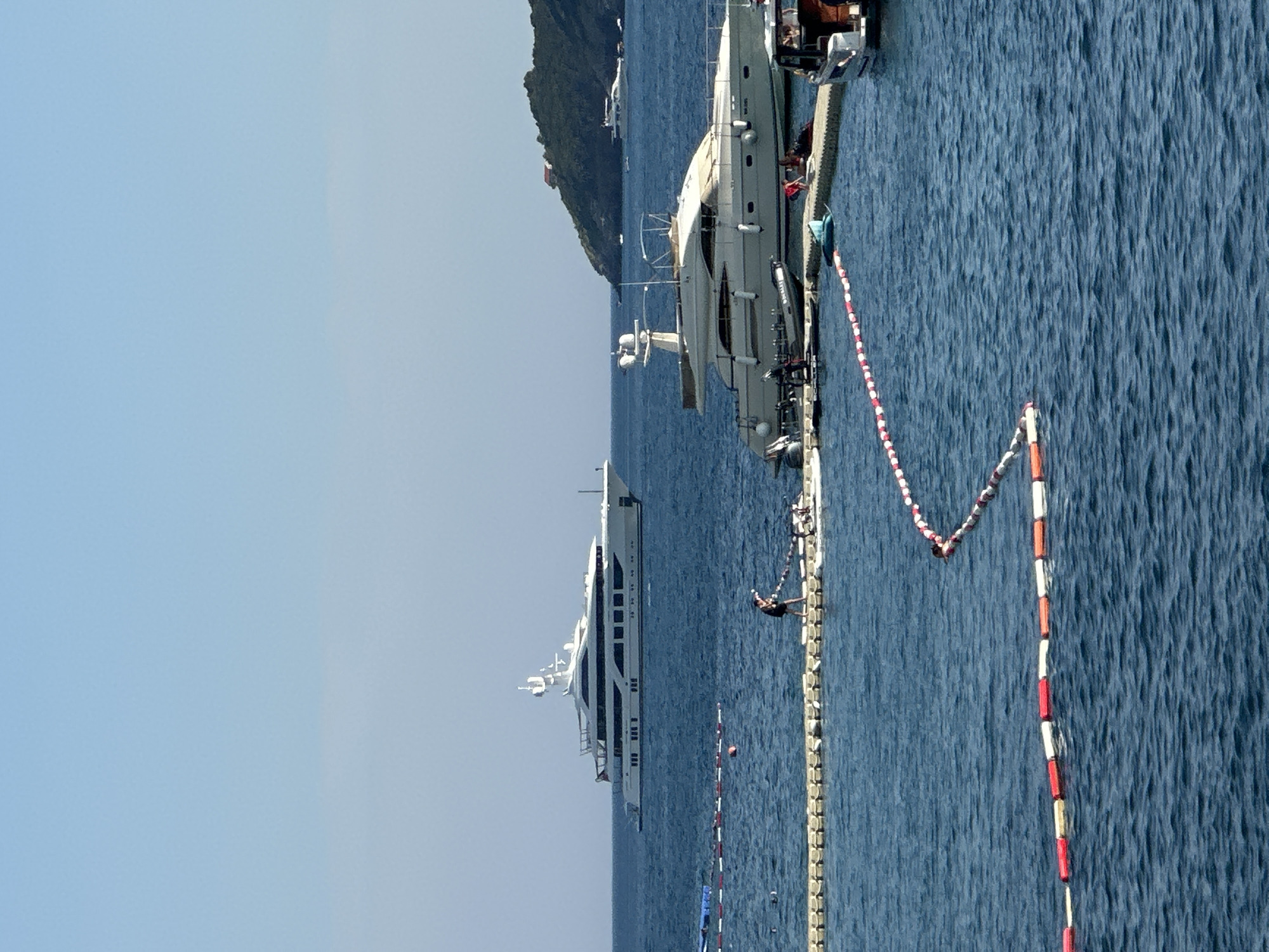 A sunny day at sea off the coast of Budva, Montenegro. Two large yachts are docked, one closer and more detailed than the other. The closer yacht is white with multiple decks and windows, and it appears to be moored to a floating dock. A person is visible on the dock, facing the water, with their back to the camera. They are wearing dark shorts and a dark top. A rope or line with red and white buoys stretches across the water, separating areas. The water is a deep blue and appears calm with slight ripples. In the background, a lush, green, tree-covered hill rises from the sea, with a hint of a building or structure on its peak. The sky is a clear, light blue with no visible clouds. The overall scene suggests a peaceful day of leisure or vacation. The image is oriented vertically.