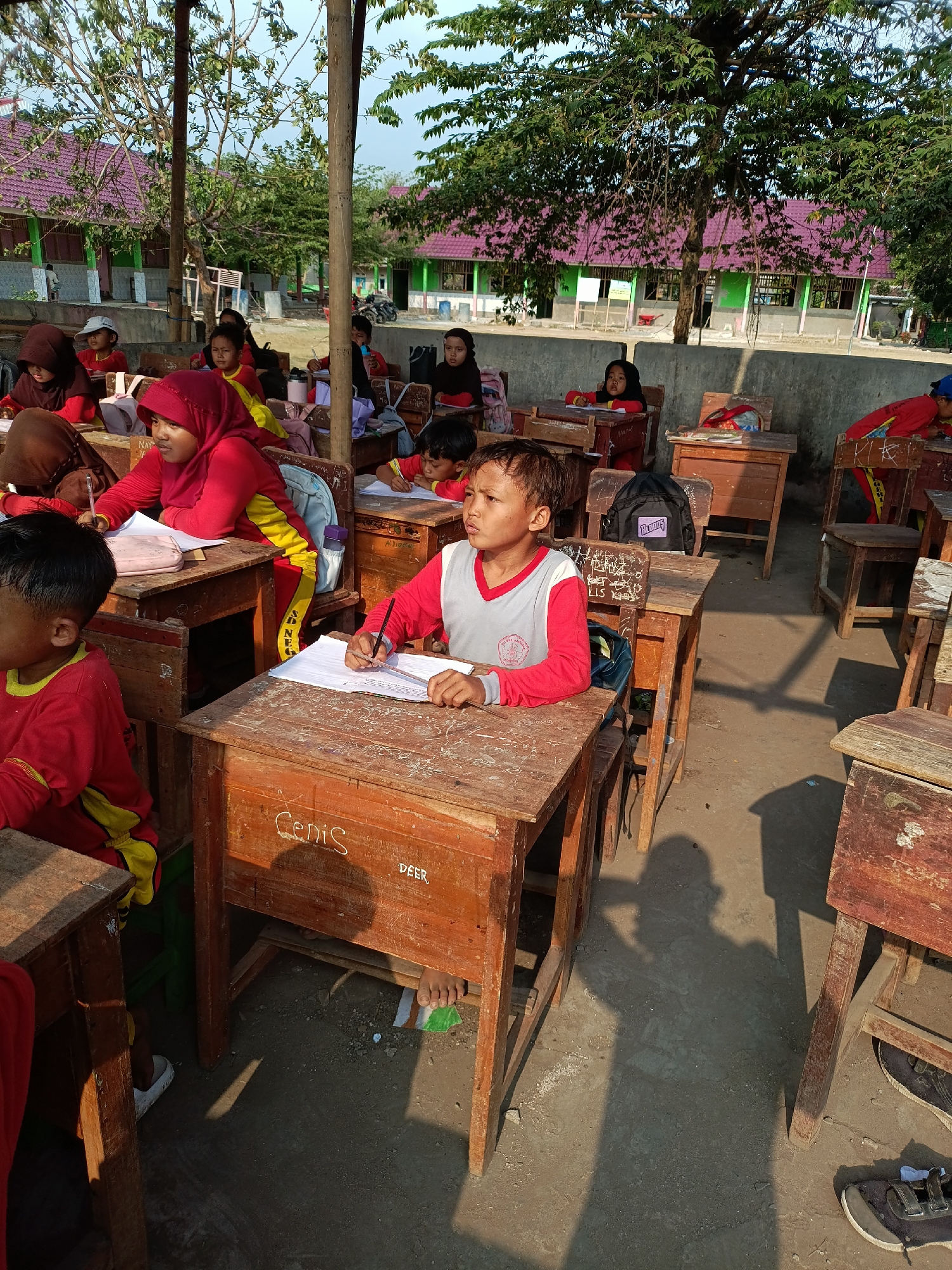 In an outdoor classroom setting in Sumberejo, Indonesia, several young students are seated at rustic wooden desks, engaged in learning. The desks are simple, open-fronted structures with attached benches. Sunlight casts strong shadows across the dirt and gravel ground. The background shows a row of school buildings with green walls and purple roofs, partially obscured by trees.
In the foreground, a boy in a red and gray school uniform is diligently writing on a piece of paper. He has a focused expression and is holding a pen. To his left, another student, also in a red uniform and headscarf, is writing. Further to the left, a student in a brown headscarf and red uniform is partially visible. In the middle ground, other students can be seen at their desks, some writing, others looking ahead. One student in a red and yellow uniform is looking off to the side. A black backpack rests on the back of a chair. The overall atmosphere suggests an informal and practical educational environment.