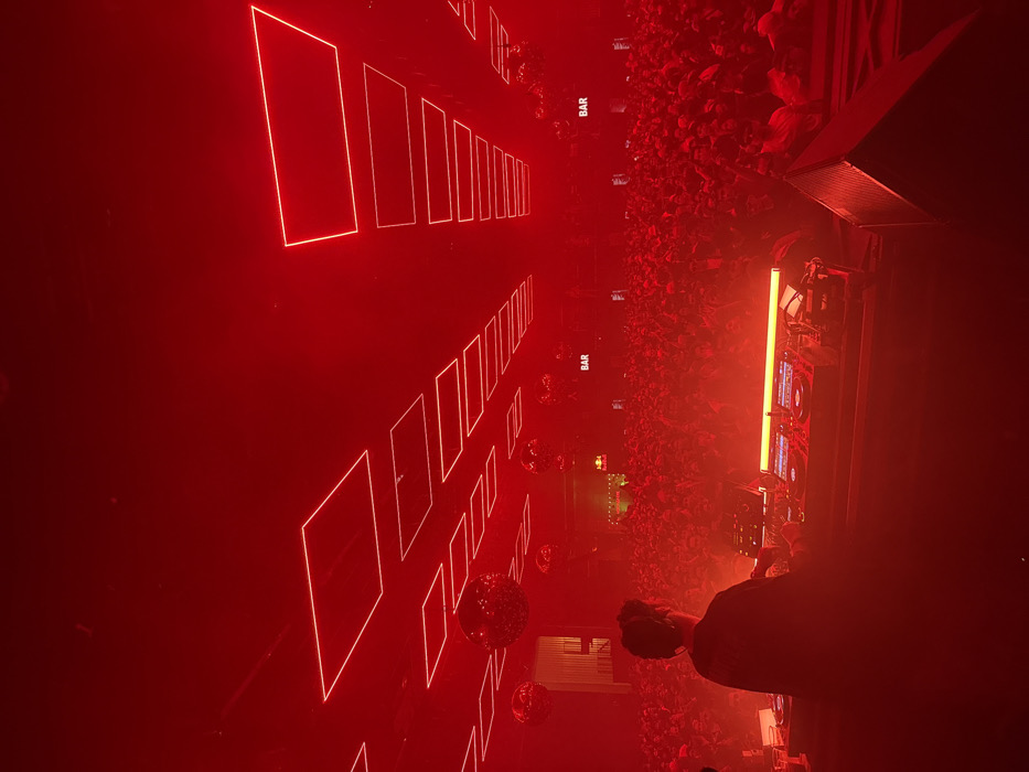 A nightclub or concert venue bathed in intense red light. A DJ station and large speakers are visible on the right, near a dense crowd. The dance floor has rows of illuminated rectangular shapes. Several disco balls are scattered in the scene. Small "BAR" signs are visible. A person with headphones is near the DJ station.