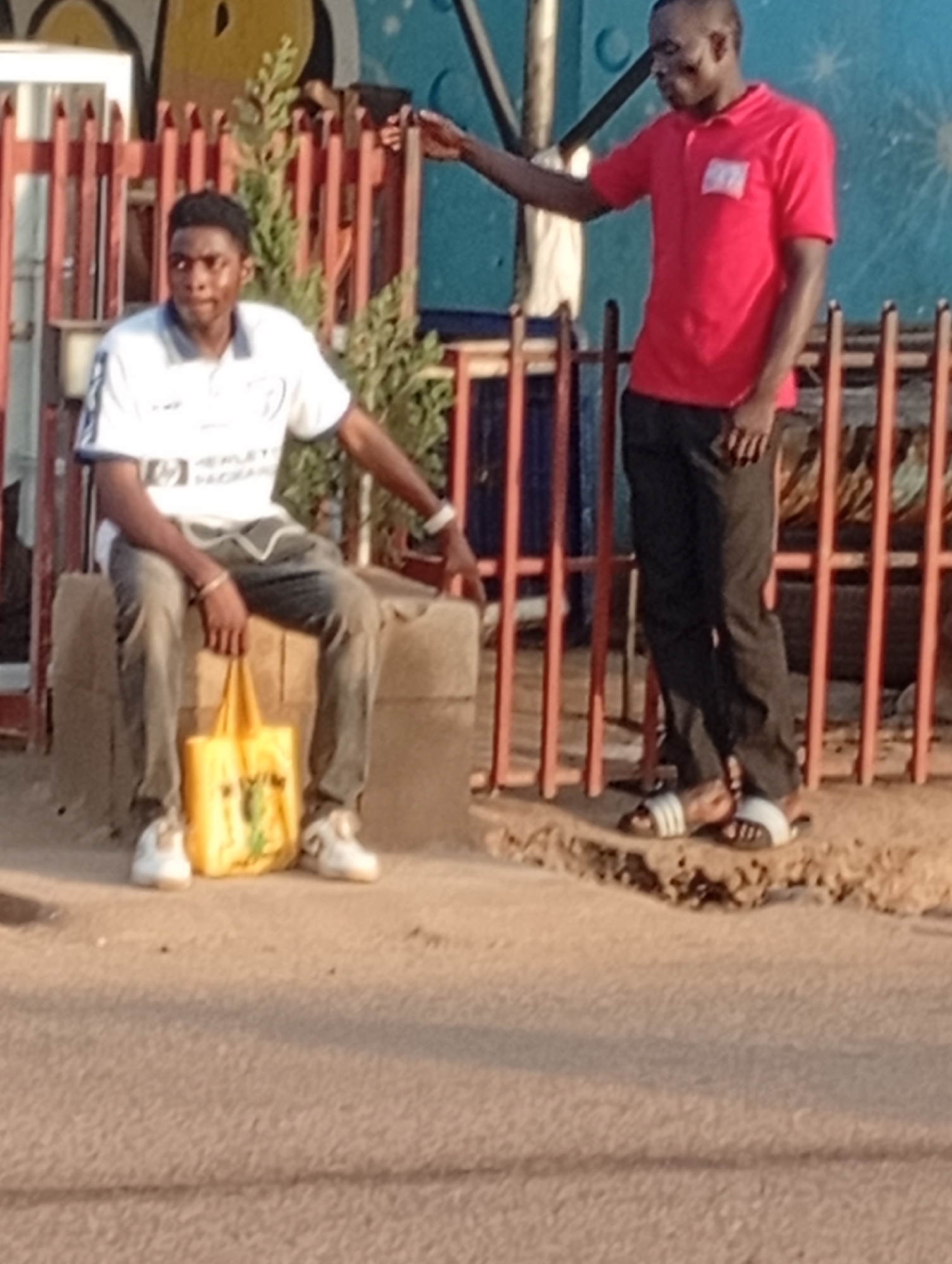 Two men are visible. One man is sitting on a concrete block holding a yellow bag. He is wearing a white t-shirt, grey pants, and white shoes. The other man is standing next to him, wearing a red shirt, black pants, and black and white sandals. They appear to be in front of a building or structure with a red metal fence. The location is identified as Narayi, Nigeria.
