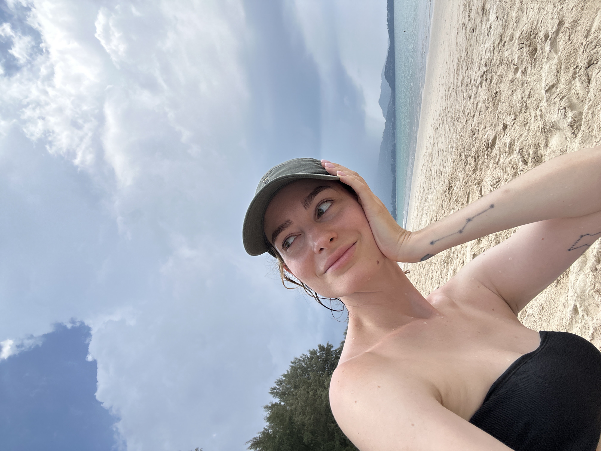 A young woman is taking a selfie on a beach near Choeng Thale, Thailand. She wears a black strapless top and a dark green cap. She is slightly wet, suggesting she may have been in the water recently. Her left hand rests on her head, showing a constellation tattoo on her forearm. The beach has light brown sand, and the sky is overcast with scattered clouds. In the background, there are trees and distant mountains.