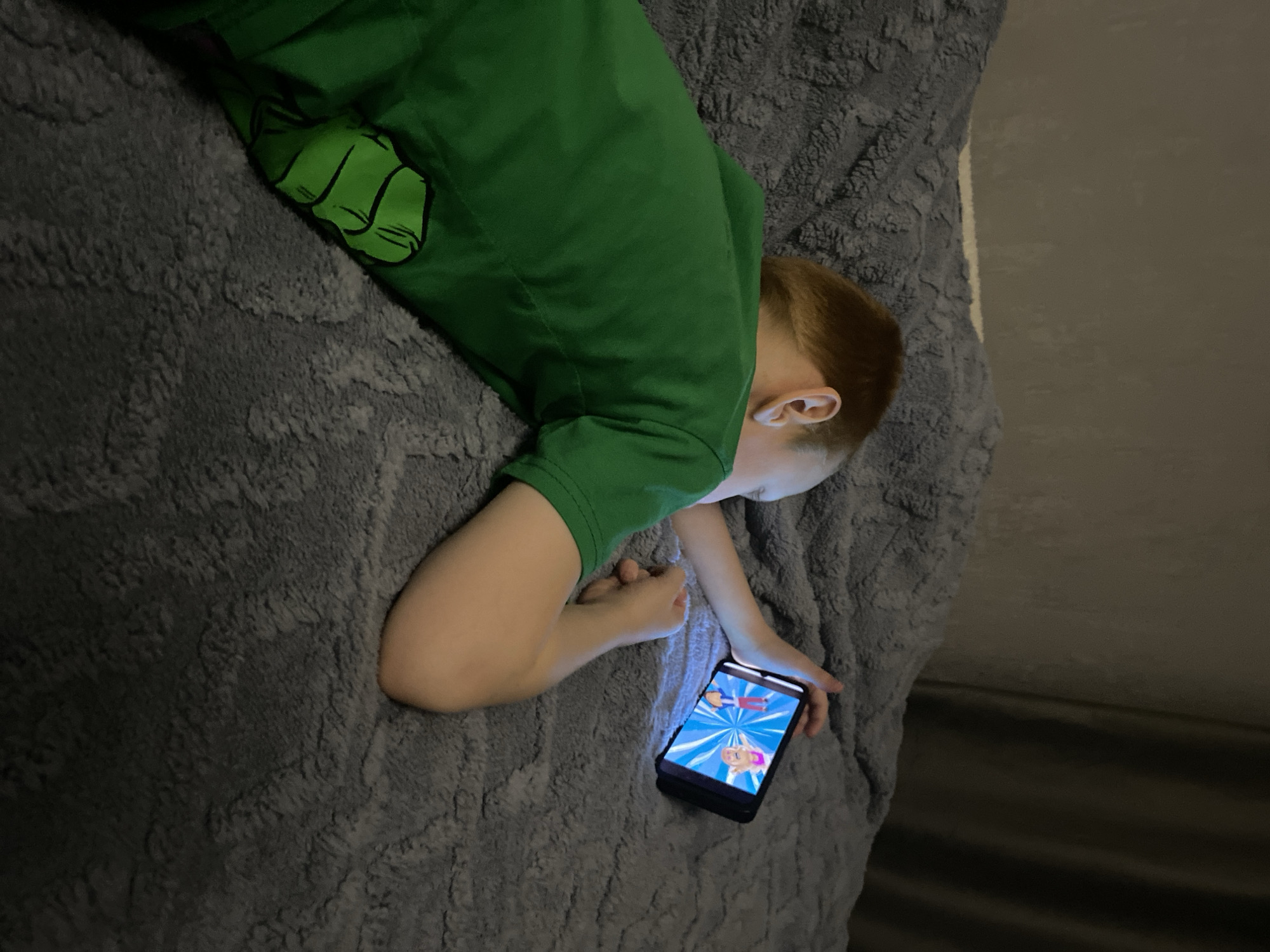 A young, light-skinned boy with red hair is lying on his stomach on a grey, textured blanket. He is wearing a green t-shirt with a cartoon graphic on the back. He is holding a black smartphone in his right hand, and the screen is illuminated, displaying a cartoon graphic. The background is a dark, undefined space. The location, Noviy, Russia, is not discernible from the image.