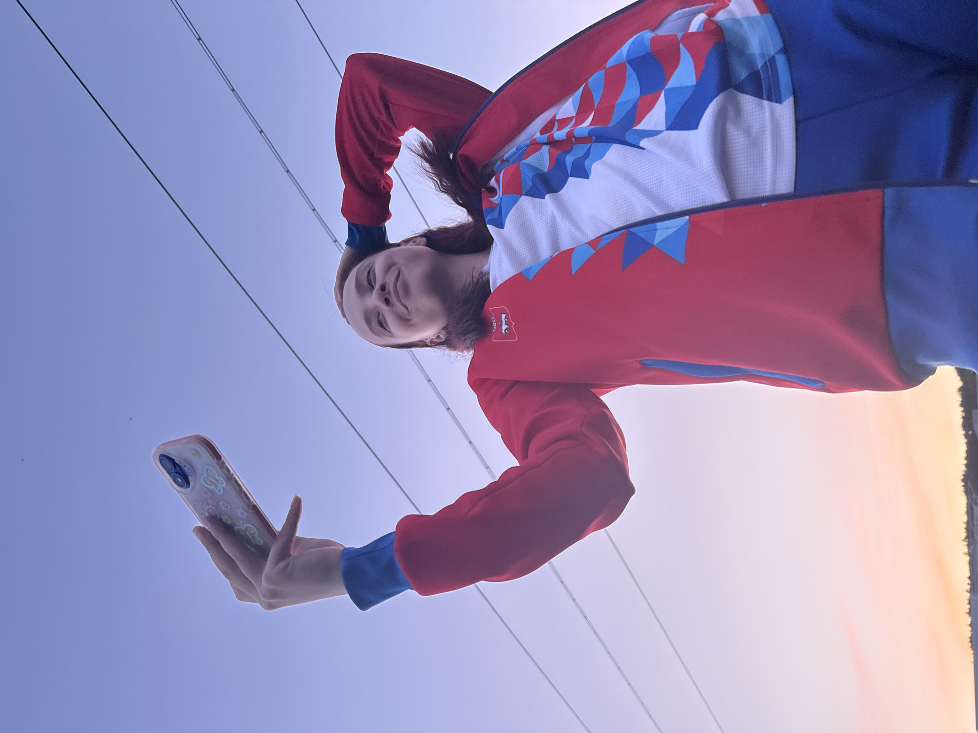 A woman is photographed from a low angle, holding a smartphone in her right hand, angled upwards towards the camera, as if taking a selfie. She's wearing a red, white, and blue jacket. Electrical power lines are visible diagonally across the upper portion of the frame, suggesting an urban or suburban setting. The sky ranges from a light blue to a pale orange at the horizon, indicating either sunrise or sunset. While the picture was taken in Khvalynsk, Russia, there is nothing within the image to prove the location; the specific location cannot be determined.