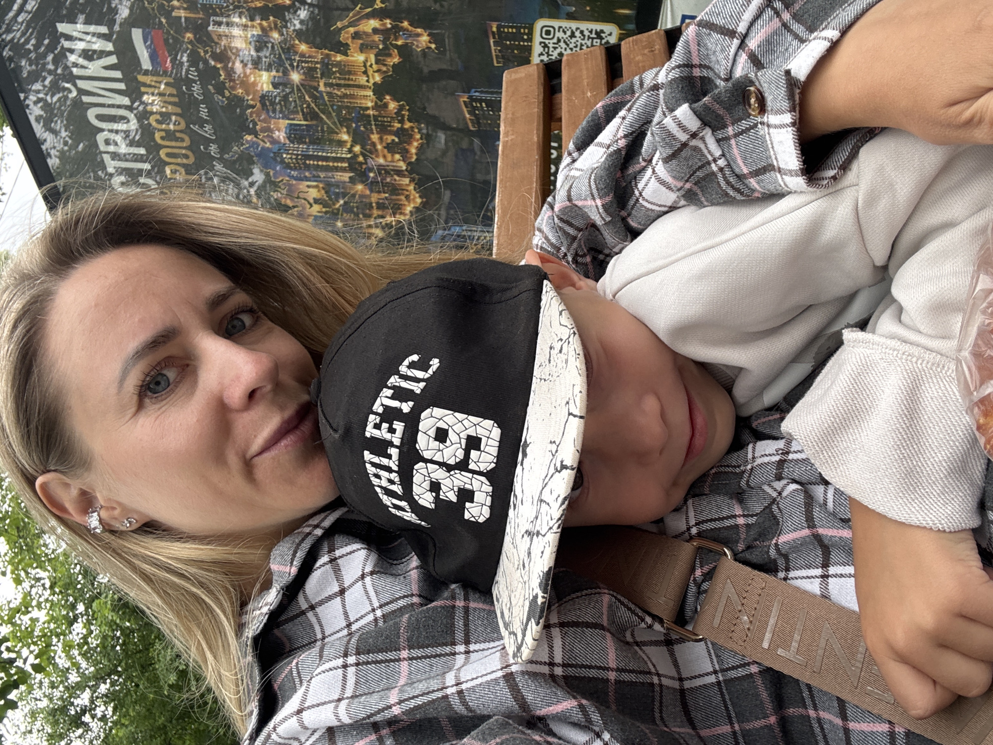 A woman and a child are seated on a wooden bench, possibly outdoors. The woman, with blonde hair and blue eyes, is wearing a plaid shirt and an ear cuff. She is holding the child, who is wearing a black baseball cap with "ATHLETIC 39" printed on it and a plaid shirt. The child appears to be leaning into the woman. In the background, there is a large banner with text in Russian and an image of a city skyline at night. A QR code is visible on the right side of the banner. The overall setting appears to be a public space within the city of Chaikovsky, Russia, given the Russian language on the banner.