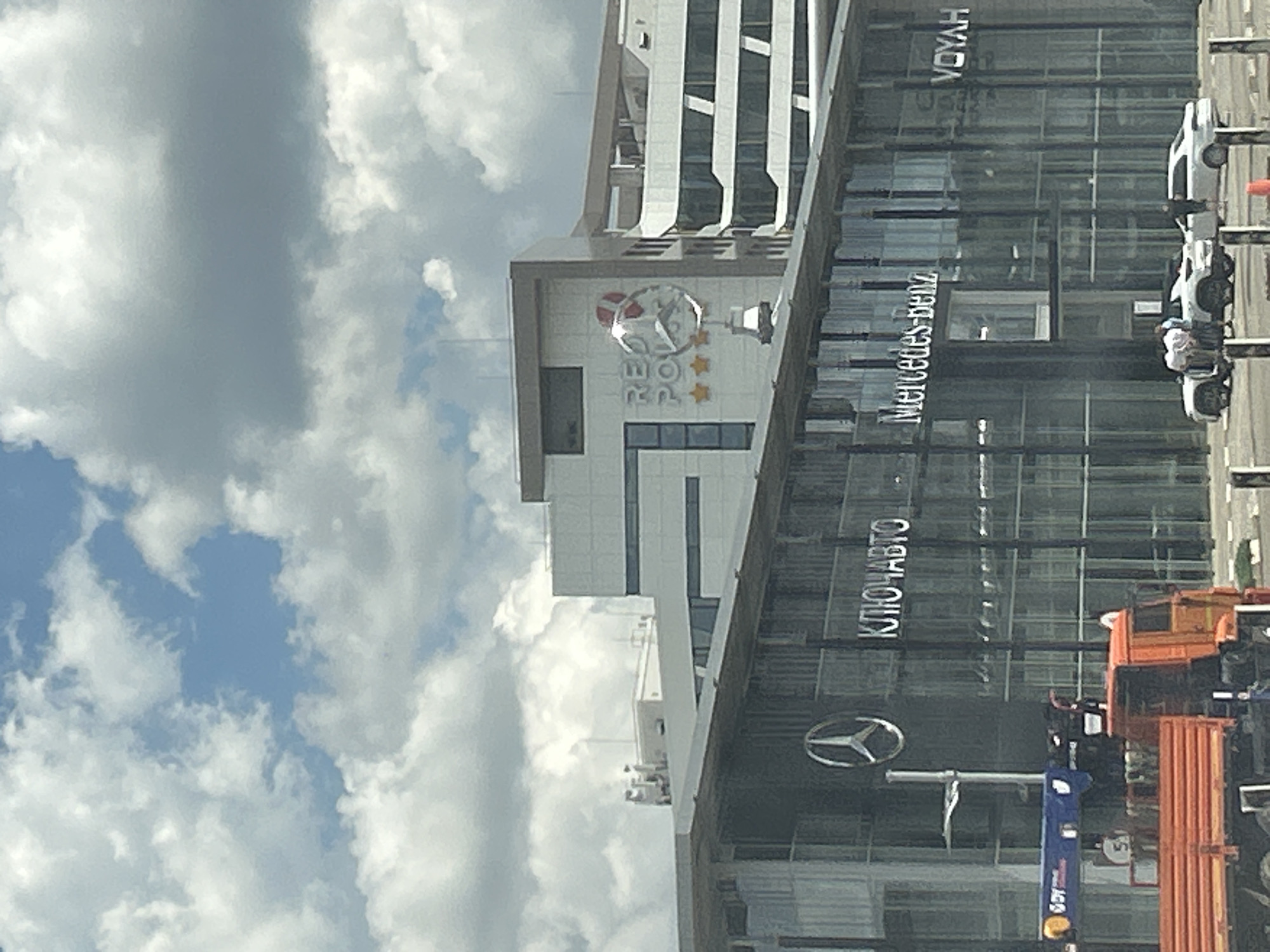 This image shows a modern building with a glass facade, likely a car dealership, under a cloudy sky. The building features large windows and illuminated signage. One prominent sign displays the Mercedes-Benz logo and text. Another sign on the building reads "КЛЮЧАВТО" in large, illuminated letters. Further up the building, there is a sign with a circular logo containing stars, possibly representing a brand or event.
Several cars are visible outside the building, including a white van and other vehicles partially obscured by the foreground elements. A person can be seen near one of the cars. In the foreground, there are construction or utility vehicles, including an orange truck with a crane-like attachment and a blue lifting device. The overall scene suggests a commercial area.
The time of day appears to be daytime, with bright lighting reflecting off the glass. The sky is filled with a mix of white and gray clouds, indicating overcast or partly cloudy weather. The text visible provides specific clues to the location and businesses present. "Mercedes-Benz" indicates a car dealership, and "КЛЮЧАВТО" is a Russian car dealership chain. The presence of what appears to be a construction site or ongoing work around the building suggests possible development or maintenance.
