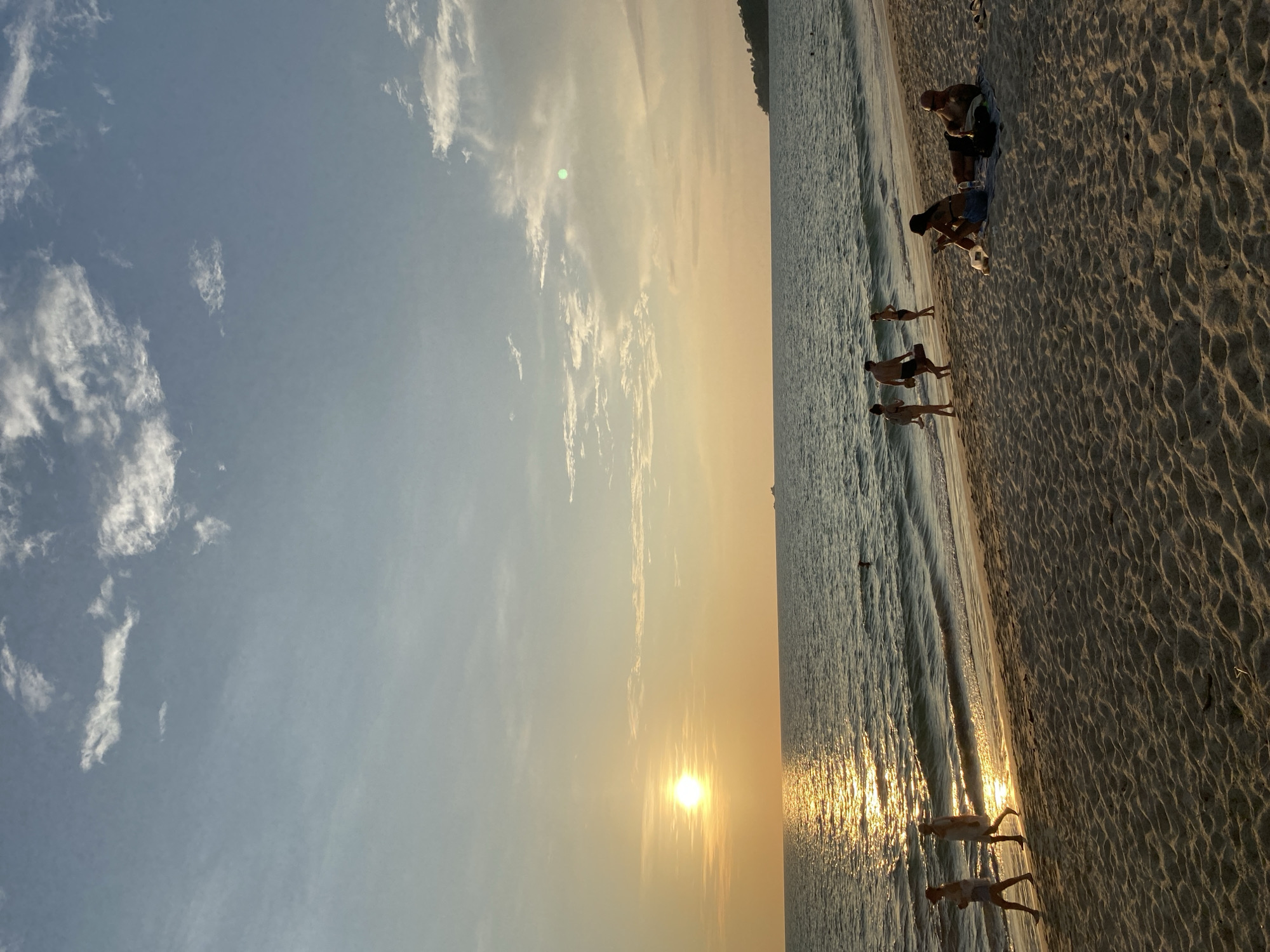 This photograph captures a serene sunset scene on a sandy beach in Choeng Thale, Thailand. The image, originally in a vertical orientation, depicts a wide expanse of the sky and sea, with a strip of beach on the right.
The time of day is clearly sunset, with the sun appearing as a bright, luminous orb positioned low on the left side of the horizon. Its golden light bathes the sky, which transitions from a soft blue at the top to warm yellow and orange hues around the sun. Scattered white, wispy clouds are visible across the sky. The sun's reflection creates a brilliant, shimmering path across the calm ocean waves, extending towards the foreground.
Several people are present on the beach and in the shallow water, enjoying the evening. On the sandy shore, which features distinct ripple patterns from the waves, two individuals are seated further from the water's edge. One person has a bare torso and wears shorts, while the other is seated nearby. Closer to the waterline, multiple figures are seen wading or standing in the shallow surf. Their forms are largely silhouetted against the bright sun reflection, suggesting activities like walking, relaxing, or observing the sunset. At least four distinct figures can be identified in and near the water, with more appearing as partial silhouettes.
The overall scene conveys a sense of peaceful leisure and natural beauty. The waves are gentle, creating a tranquil atmosphere. There is no visible text in the image.