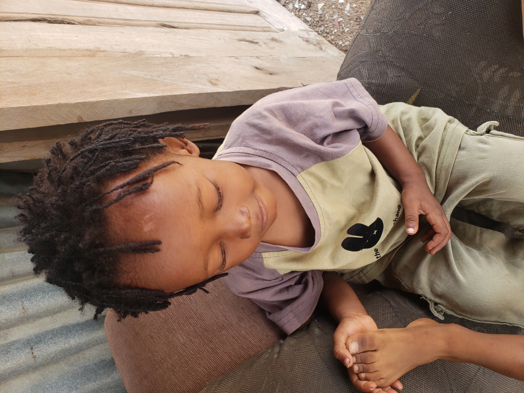 A young child with dark skin and dreadlocks is captured lying down, likely resting or sleeping, in an outdoor setting in Adankolo, Nigeria. The child's eyes are closed, and they have a peaceful expression. They are dressed in a two-toned t-shirt, featuring a light purple upper portion and a light olive green lower section, paired with light olive green pants. A black graphic, resembling a butterfly or an abstract shape, is visible on the olive green part of the shirt, along with partial text that reads "the shang".
The child is reclining on a dark, textured fabric surface, possibly a mat or cushion. In the background, several light-colored wooden planks or boards are visible, suggesting a construction area or a rustic environment. Scattered debris, appearing as small stones or broken fragments, lies on the ground near the wooden structures. A hint of corrugated metal can be seen in the lower left corner of the frame. The scene is illuminated by natural daylight, indicating it is daytime. The overall impression is one of serene rest.