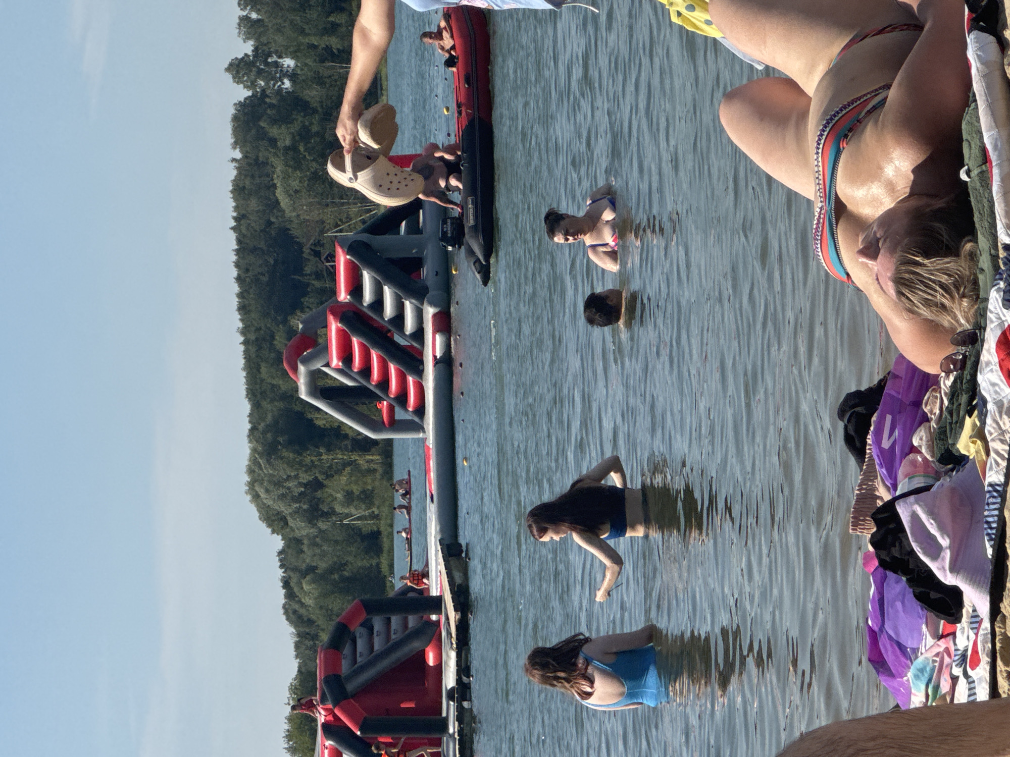 A group of people are at a lake with inflatable water attractions in Kraskovo, Russia. The sky is clear and blue. In the foreground, a woman in a colorful bikini lies on a towel on the beach, facing away from the camera. A pile of clothes and accessories is on the sand next to her. In the water, two women are standing. One woman wears a blue swimsuit, and the other wears a blue bikini. Further out in the water, a man is visible in a boat, and another person is partially submerged. In the background, large inflatable structures are set up on the water. Several other people can be seen in and around the water attractions. The lake is surrounded by a dense green forest.