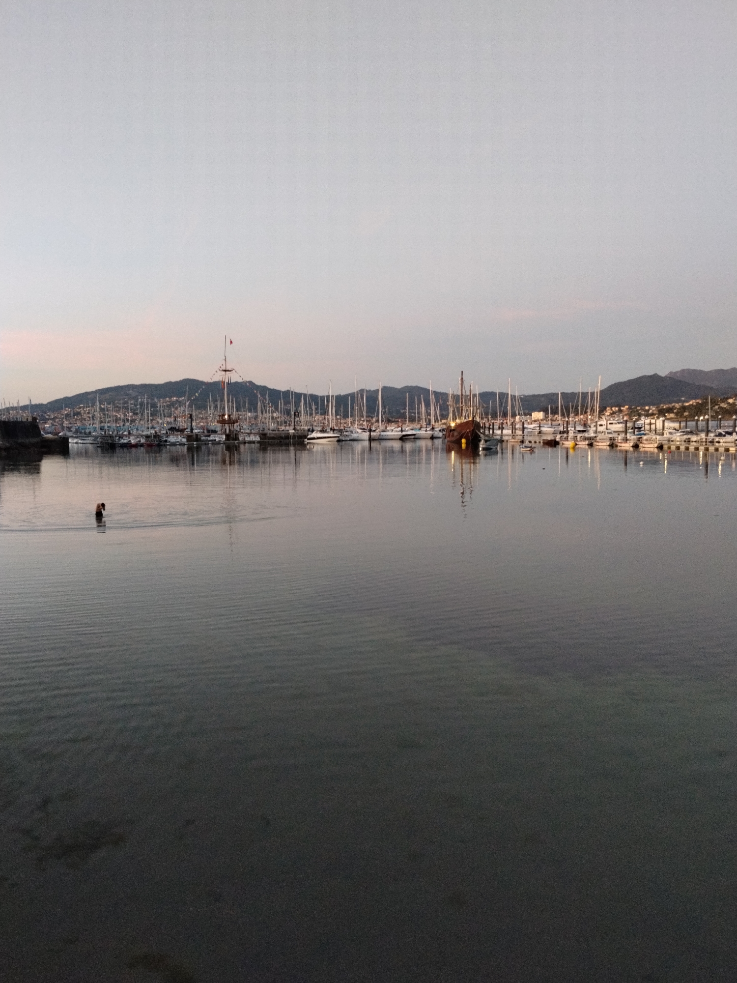 The image captures a serene marina scene in Baiona, Spain, during twilight. In the foreground, the calm water's surface is rippled by gentle waves. A solitary figure, appearing to be a person, is wading in the water near the left side of the frame. Behind this figure, the marina is lined with numerous sailboats and yachts, their masts creating a dense forest of vertical lines against the backdrop of a hillside town. A distinctive tall ship with a wooden hull and multiple masts stands out among the modern vessels. The sky above is a pale gradient of light blue and pink, indicating the end of the day. The overall atmosphere is tranquil, with the soft light reflecting on the water and the distant lights of the town twinkling on the hillside.