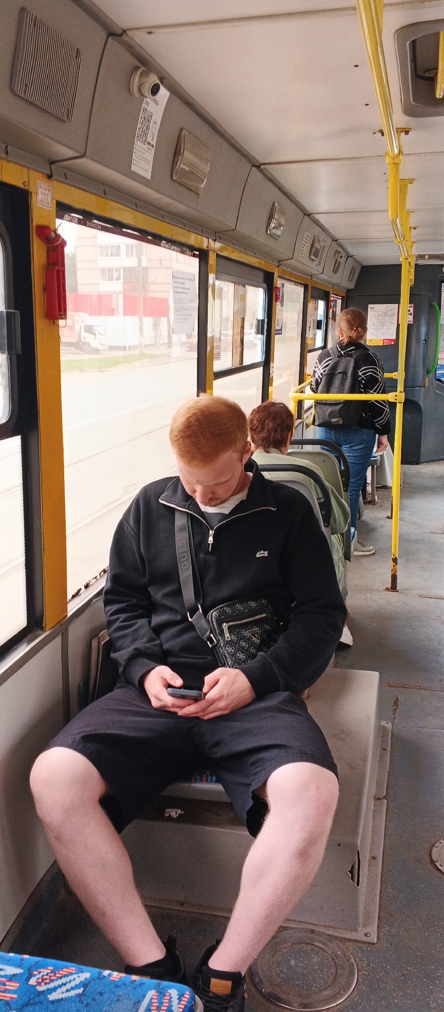 The image depicts the interior of a public transport vehicle, likely a bus or trolleybus, within the city of Izhevsk, Russia. The primary subject is a young man with red hair, seated and looking down at his mobile phone. He is wearing a black zip-up sweatshirt, black shorts, and black sneakers. He has a black crossbody bag with a patterned design slung over his shoulder.
In the background, other passengers are visible. To the right, a woman with blonde hair pulled back in a ponytail is standing and facing away from the camera. She is wearing a black and white patterned jacket and blue jeans, and carrying a backpack. Further ahead, another passenger, with short, dark curly hair, is seated.
The interior of the vehicle is characterized by grey paneling, yellow grab poles, and large windows that offer a view of the outside. The exterior view shows buildings and a white truck. The seats appear to be made of a grey, worn material. The overall lighting suggests daytime. The presence of a QR code sticker and signage on the interior panels indicates this is a functional public transport environment.