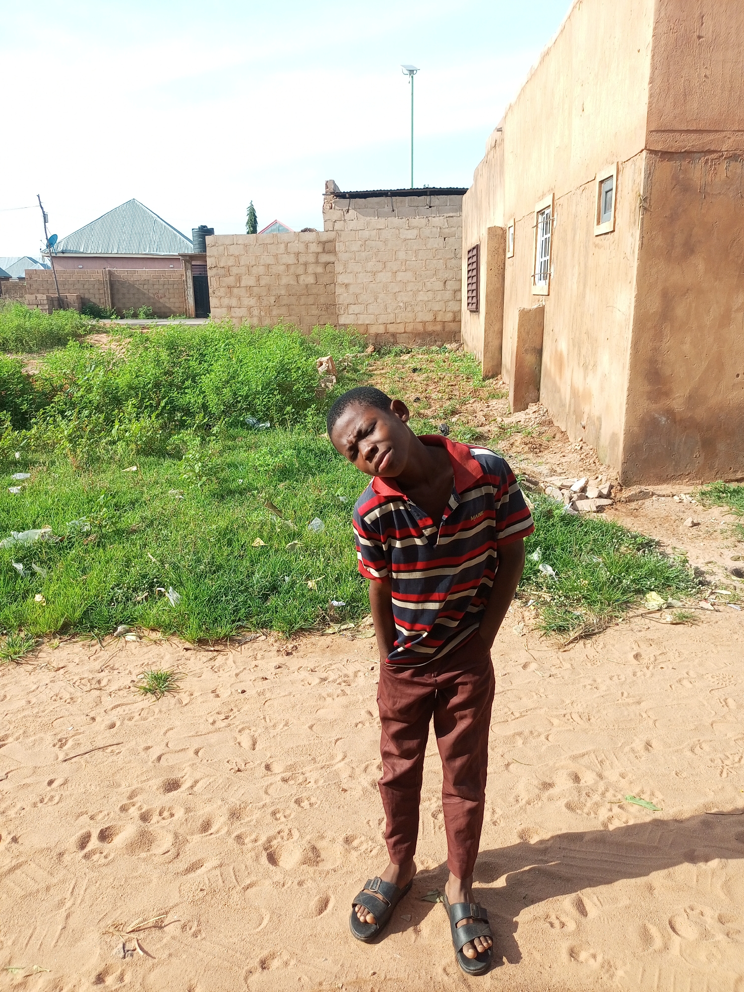 A young boy stands in a sandy, outdoor area with buildings and vegetation in the background. The boy is wearing a striped polo shirt and brown pants, with his hands in his pockets. He is looking towards the camera with his head tilted. The environment appears to be a somewhat unkempt area with scattered debris and dry grass. The buildings suggest a residential or developing area. The presence of foot prints in the sand indicates activity in the area.