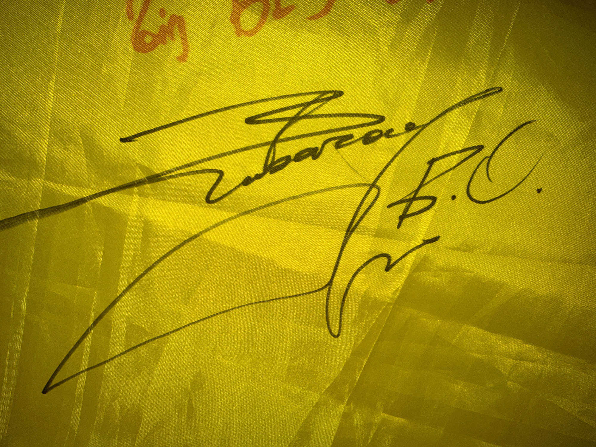 The image displays a close-up, angled shot of a signature in black ink on a bright yellow, textured fabric, likely silk or satin, which exhibits a lustrous sheen and visible weave. The fabric is creased and folded, creating shadows and highlighting the texture. The signature appears to be that of a person, with a flourish and legible parts including what looks like "Robarzo" and possibly an initial "B.C." or "B.G." underneath. To the right of the signature, faint orange markings are visible on the fabric, possibly a logo or another decorative element. The image is sharply focused on the signature and fabric. The scene does not provide any specific geographical indicators for Aalsmeer, Netherlands.