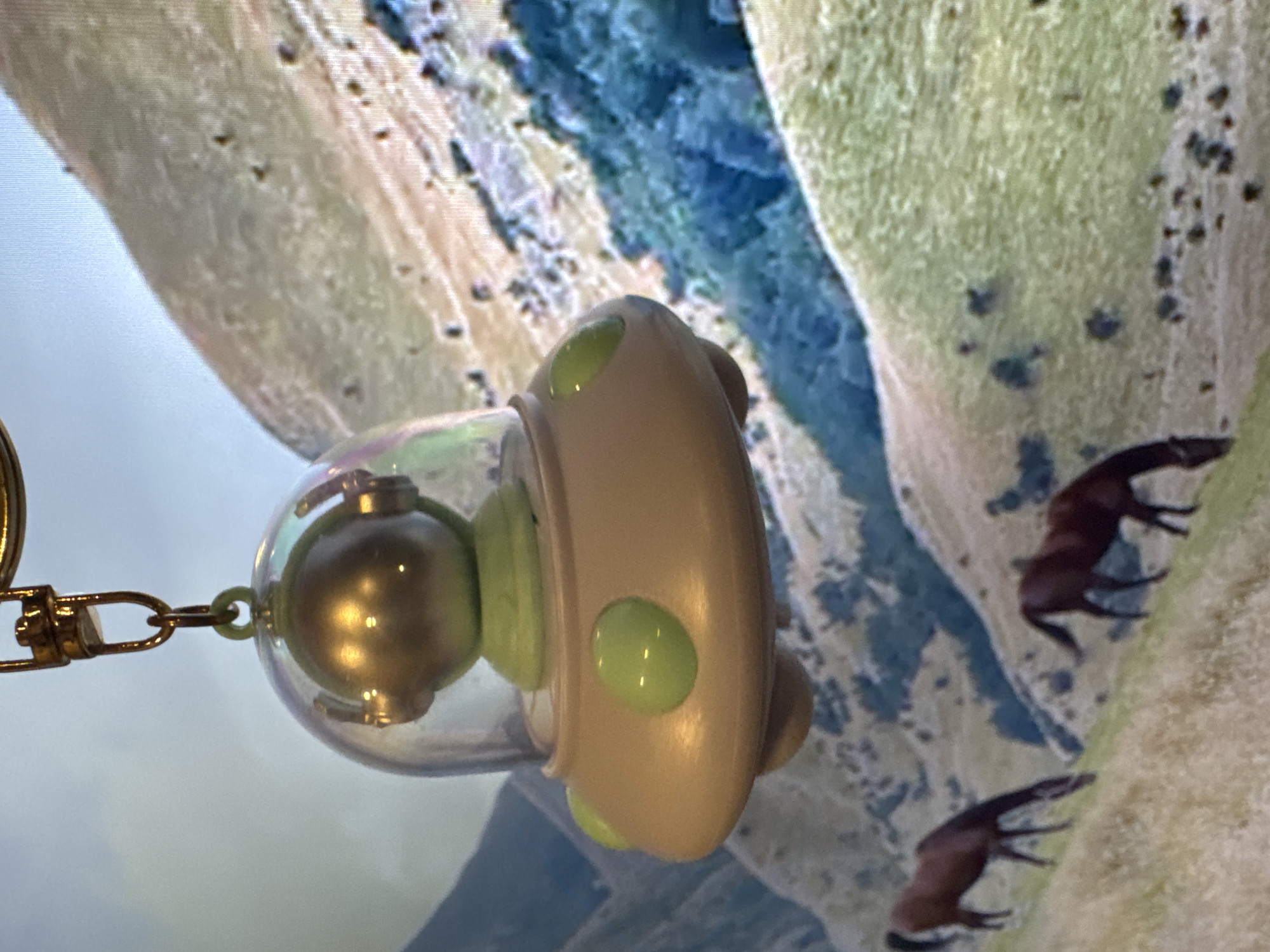 An angled, close-up photograph features a decorative keychain or small toy in the foreground, set against a background displaying a landscape. The main object is oriented horizontally, appearing suspended, and consists of a light beige or tan body with several glossy, light green circular accents. A clear spherical dome protrudes from one end, encasing a golden metallic sphere and a light green base. A golden metal chain and clasp are attached to the clear dome's top.
The background, which appears to be a digital screen or a large print, depicts a rugged, hilly landscape with varying shades of green grass and bluish-grey rock formations or water bodies. Two dark brown, horse-like figures are visible on the green slopes in the background, appearing to be in motion. The foreground object is in sharp focus, while the background is slightly blurred, consistent with a shallow depth of field. The overall scene is brightly lit, with reflections on the object's metallic and glossy surfaces. There is no visible text in the image. The image was captured in Bangkok, Thailand.