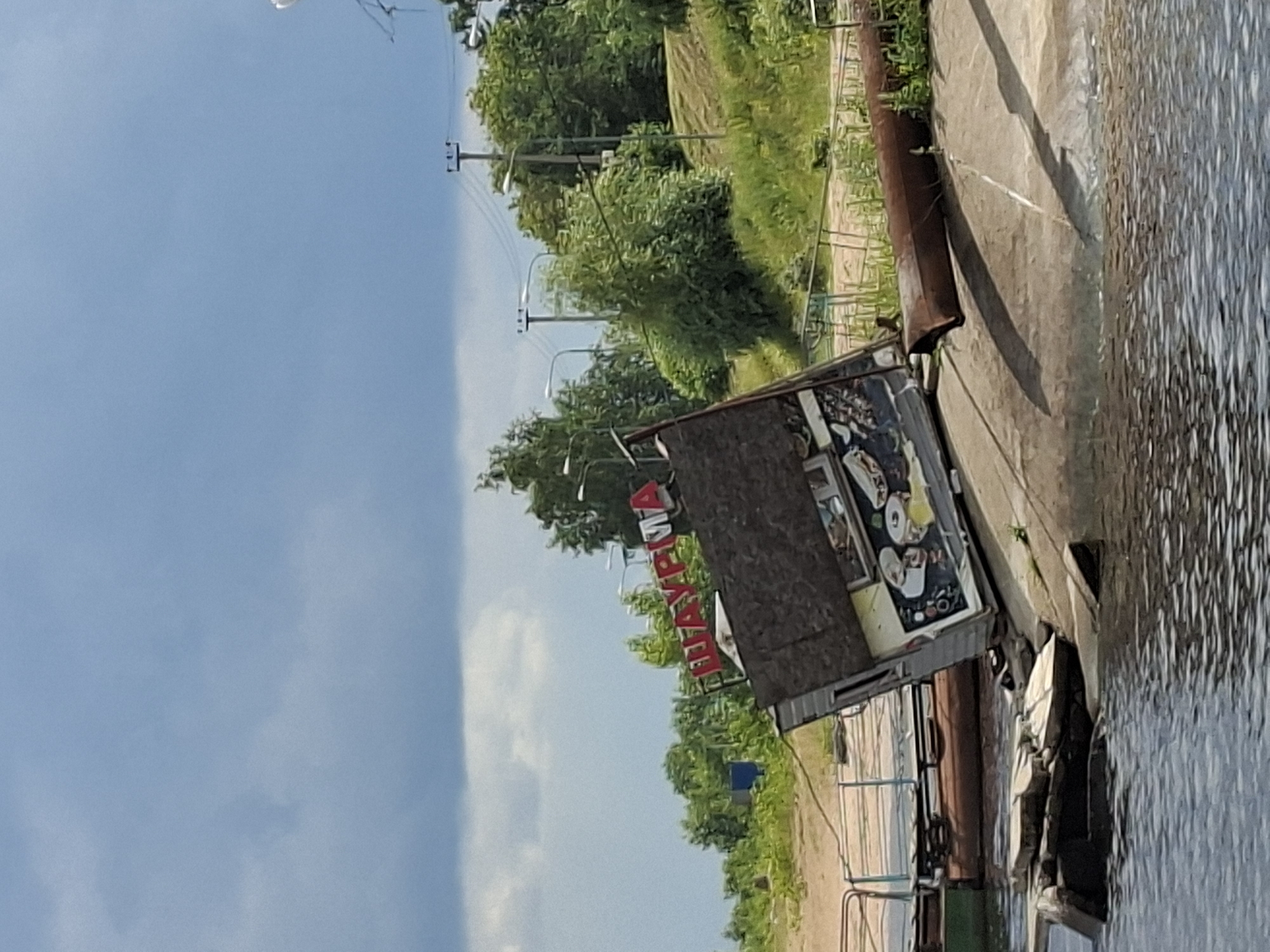 In the city of Verhnij Uslon, Russia, a dilapidated kiosk, seemingly a food stand with the Cyrillic word "ШАУРМА" (SHAURMA) prominently displayed in red lettering, is tilted precariously on a concrete embankment. The structure appears abandoned, with its wooden facade showing signs of wear and tear, and a graphic depiction of food items visible on its side. The kiosk is situated near the water's edge, with debris, including what appear to be concrete blocks and a rusty metal pipe, scattered around its base. The surrounding environment features lush green trees and vegetation, with a cloudy sky overhead. Electrical poles and wires are visible in the background, suggesting the presence of a developed area. The scene conveys a sense of neglect and disrepair.