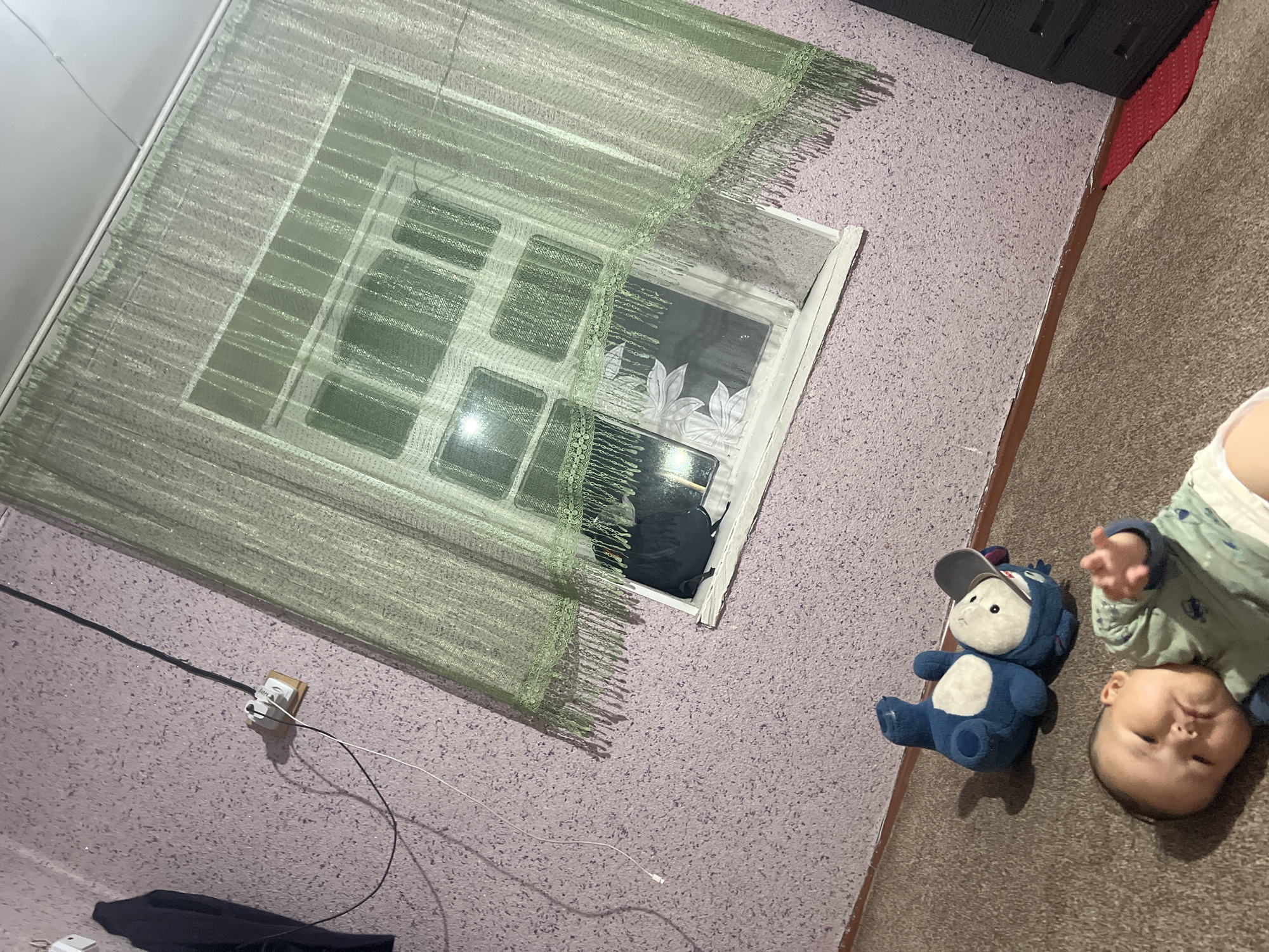 The image shows an indoor scene in Kara-Dıykan, Kyrgyzstan. The room appears to be a bedroom or living area, with a light-colored, textured wall and a carpeted floor. A window is visible on the left side of the image, covered by a sheer green curtain with a fringed hem. The window has a white frame and a decorative pattern on the glass, resembling leaves.
In the foreground, a baby is lying on its back on the carpet, looking towards the camera with a playful expression. The baby is wearing a light blue and green outfit. Next to the baby is a blue stuffed animal resembling a monster wearing a grey baseball cap.
On the wall, a power outlet with a black and white cord is visible, suggesting the presence of electrical devices. A dark piece of fabric, possibly clothing, is on the floor near the wall. A dark, tall cabinet is partially visible in the upper right corner of the image, along with a section of a red rug.
The lighting suggests it is daytime, possibly late afternoon or early evening, given the shadows. The overall mood is relaxed and domestic, with the baby and toy creating a sense of playfulness. No visible text is present in the image.