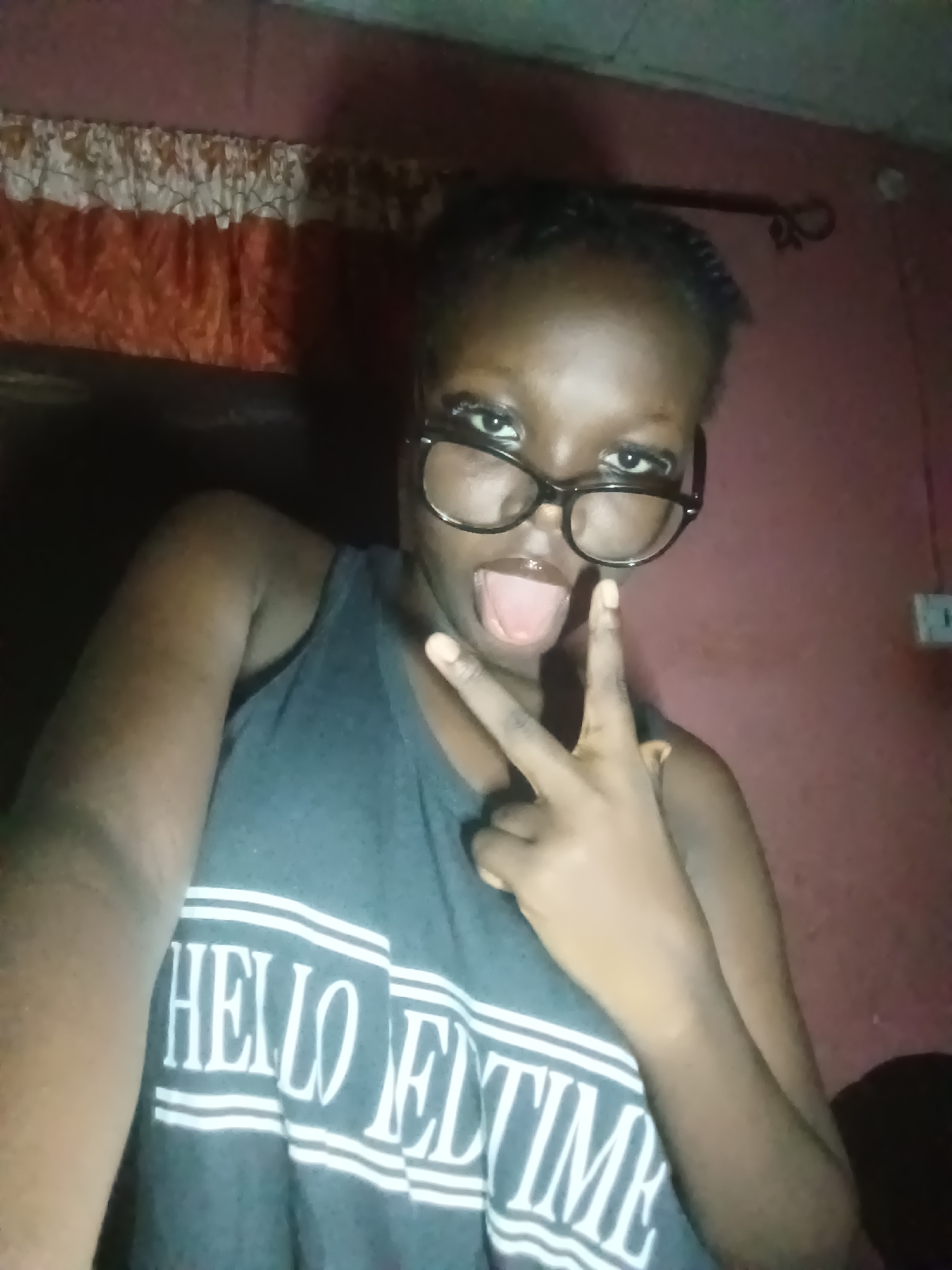 A young individual with dark skin, wearing black-framed glasses and braided hair, is depicted in a playful selfie taken indoors, likely in Onitsha, Nigeria. The person is sticking out their tongue and making a peace sign (V-sign) with their right hand, indicating a casual and expressive mood. They are wearing a dark grey tank top with prominent white text that reads "HELLO BEDTIME" across the chest.
The background suggests a dim indoor setting, likely in the evening or at night. A patterned curtain with a white and orange design is visible at the top left, above a reddish-brown wall. To the right of the person's head, a white electrical outlet or switch is mounted on the wall. The lighting is low, contributing to a cozy or private atmosphere.