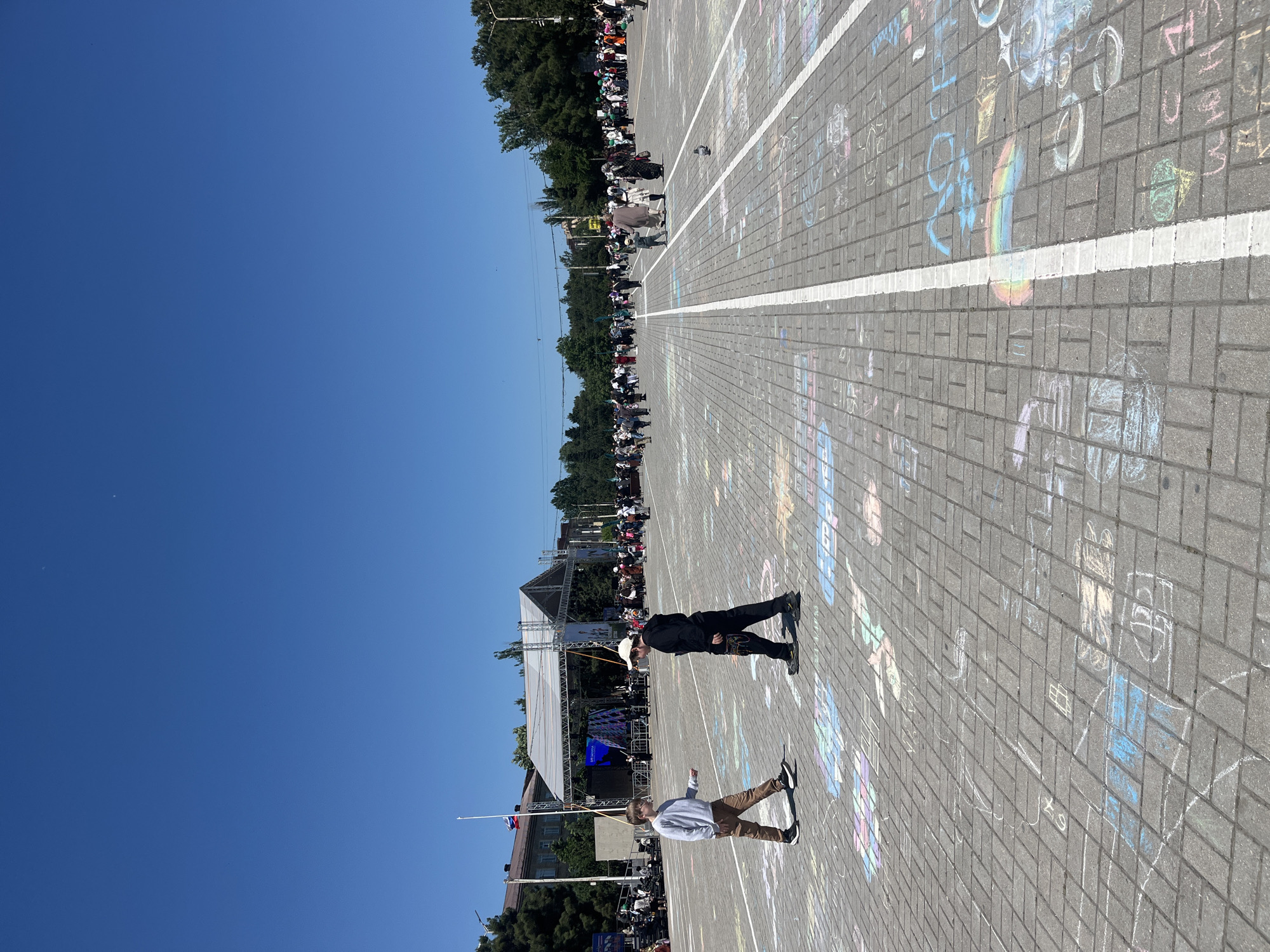 Two people stand on a brick-paved surface, marked with chalk drawings and white lines. A stage structure is visible in the background, along with a crowd of people. A Russian flag is also present. The scene is located in the city of Kaspiysk, Russia.