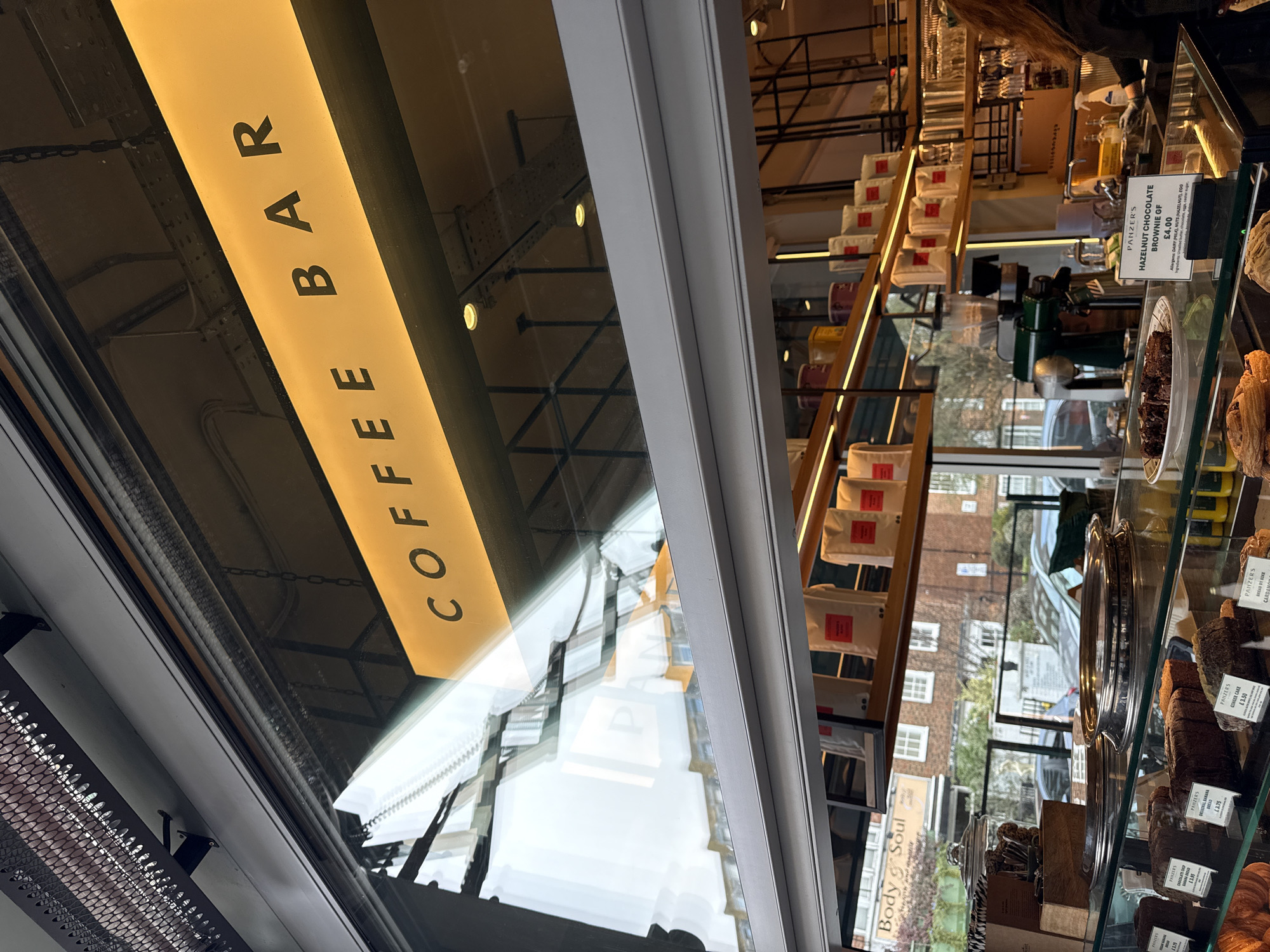 The image captures the interior of Panzers Deli and Coffee Bar, located in St John's Wood, London, United Kingdom, viewed looking outwards towards a city street. In the immediate foreground, a glass display case presents an assortment of baked goods, including various loaves of cake (e.g., "Original Banana Bread," "Ginger Cake"), croissants, and "Hazelnut Chocolate Brownie GF," each labeled with a price. Behind this display, the upper back and head of a person, identified as a staff member with long, dark hair wearing a dark garment, are visible near a stainless steel sink and coffee preparation equipment. Above, an illuminated sign reading "COFFEE BAR" is prominently displayed. Interior shelves are stocked with numerous white bags bearing red labels, presumably coffee or retail products. Through the large window, reflections reveal the exterior environment: multiple residential buildings with distinct window patterns, parked vehicles, bare trees, and a partial reflection of a "Body & Soul" sign. An overhead infrared heater with a black mesh cage is mounted above the exterior of the window frame.