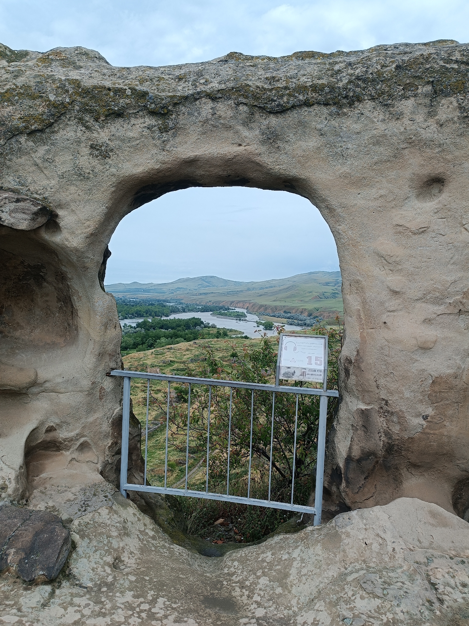 The image shows a natural rock archway in the ancient cave city of Uplistsikhe, Georgia. Through the arch, a scenic view of the Mtkvari River and surrounding rolling hills unfolds under an overcast sky. A metal railing and a small informational sign are visible in the foreground, positioned just outside the arch. The rock formations are weathered and textured, indicating their age. No people are present in the scene. The environment is natural and historical, characterized by rock structures, vegetation, and a river valley.