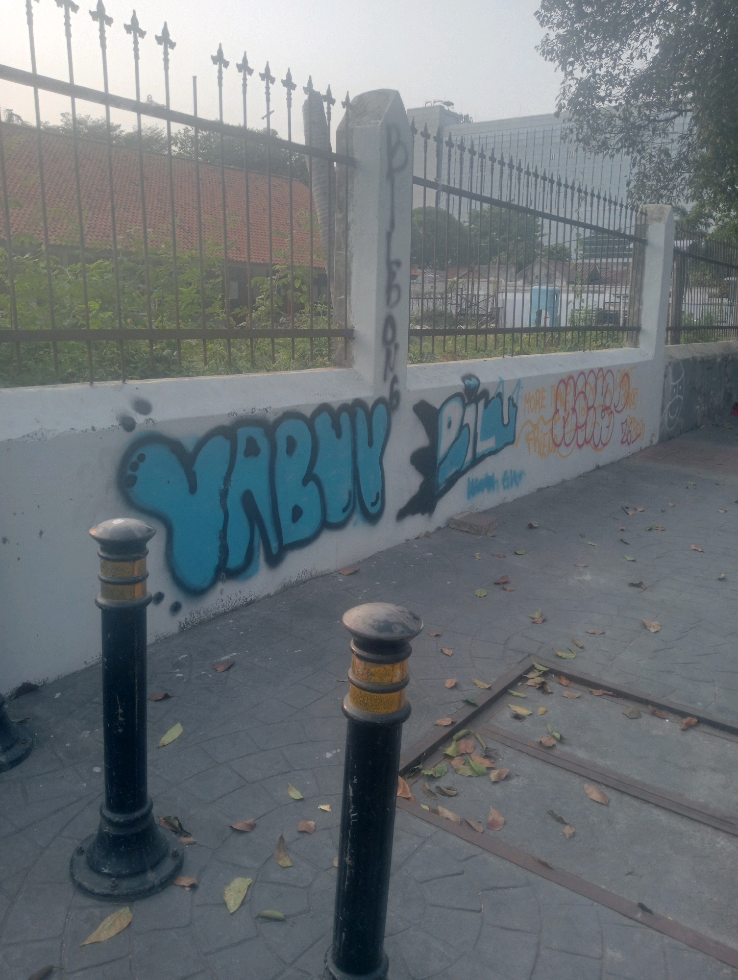 The image captures a street scene in East Jakarta, Indonesia, likely in an urban or semi-urban setting. A low white concrete wall, partially covered in graffiti, dominates the lower half of the frame. The graffiti features bold, stylized lettering in blue, black, and orange. To the right of the blue graffiti, there is orange graffiti that appears to be a tag. The wall is topped with a black metal fence with decorative finials, beyond which are trees and a building. In the foreground, two black bollards with yellow banding are positioned on a paved sidewalk. Fallen leaves are scattered across the pavement. The sky is overcast, suggesting a hazy or foggy atmosphere. The overall impression is of an outdoor public space with signs of urban decay and artistic expression.
