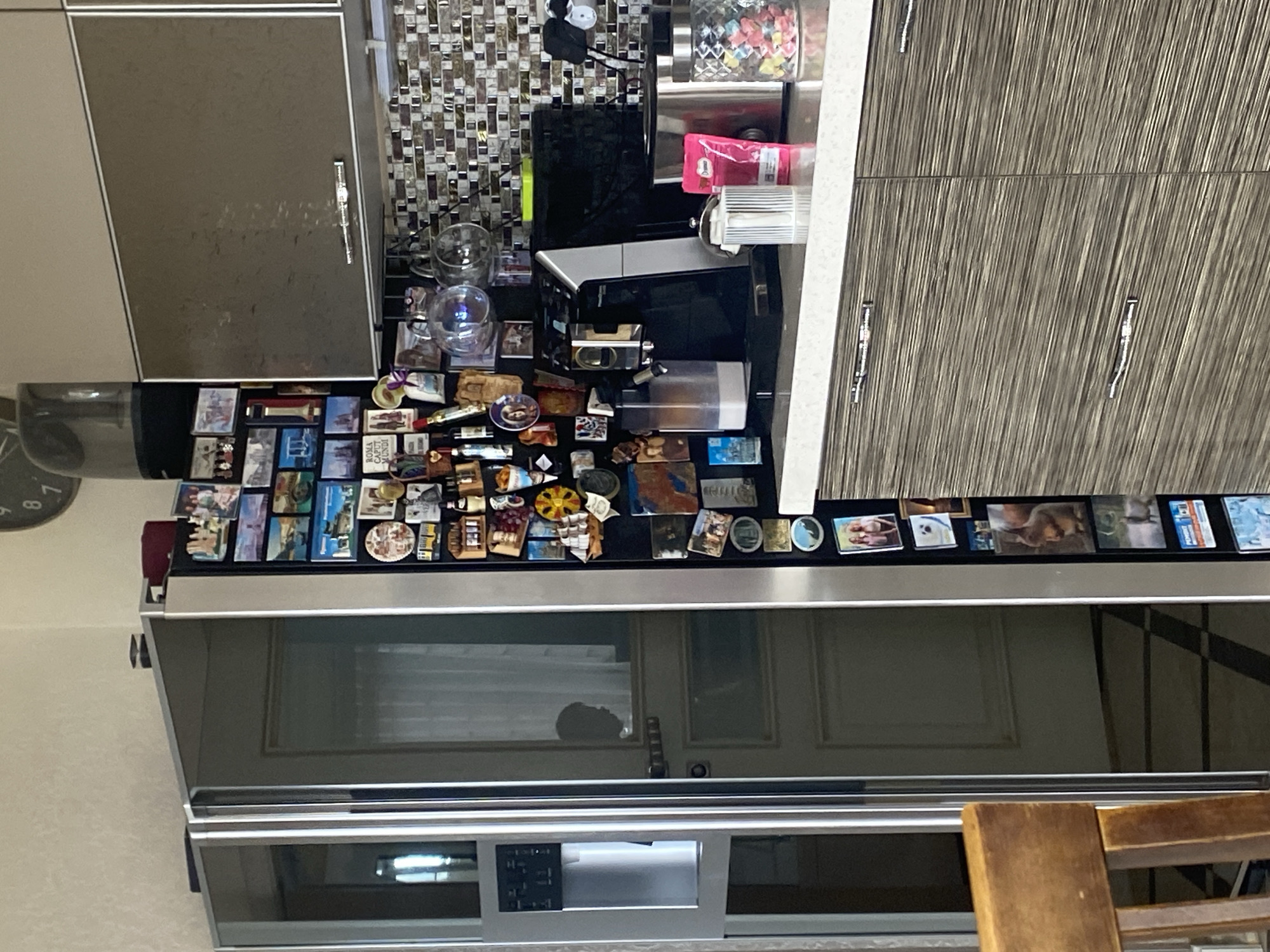 The image captures a close-up, eye-level view of a kitchen scene. A predominantly silver and dark grey color palette dominates the space, with accents of wood grain on cabinetry. On the left, a tall, sleek refrigerator is covered with numerous small magnets, primarily depicting travel destinations and various images. One magnet reads "ROMA CAPUT MUNDI." To the right of the refrigerator, a dark grey countertop features several kitchen appliances, including a black coffee maker with a water reservoir, a silver toaster, and a glass jar filled with colorful, small objects, possibly candies. A bright pink bag of coffee is also visible on the counter. Above the countertop, a backsplash composed of small, rectangular mosaic tiles in shades of brown, beige, and white adds texture to the wall. A set of dark grey cabinets with silver handles is mounted above the backsplash. In the background, a glimpse of a wall clock with Roman numerals can be seen. The overall impression is a modern, functional kitchen, likely in a residential setting, devoid of any explicit indicators of its specific location in Sochi, Russia. There are no individuals present or interacting in the scene.