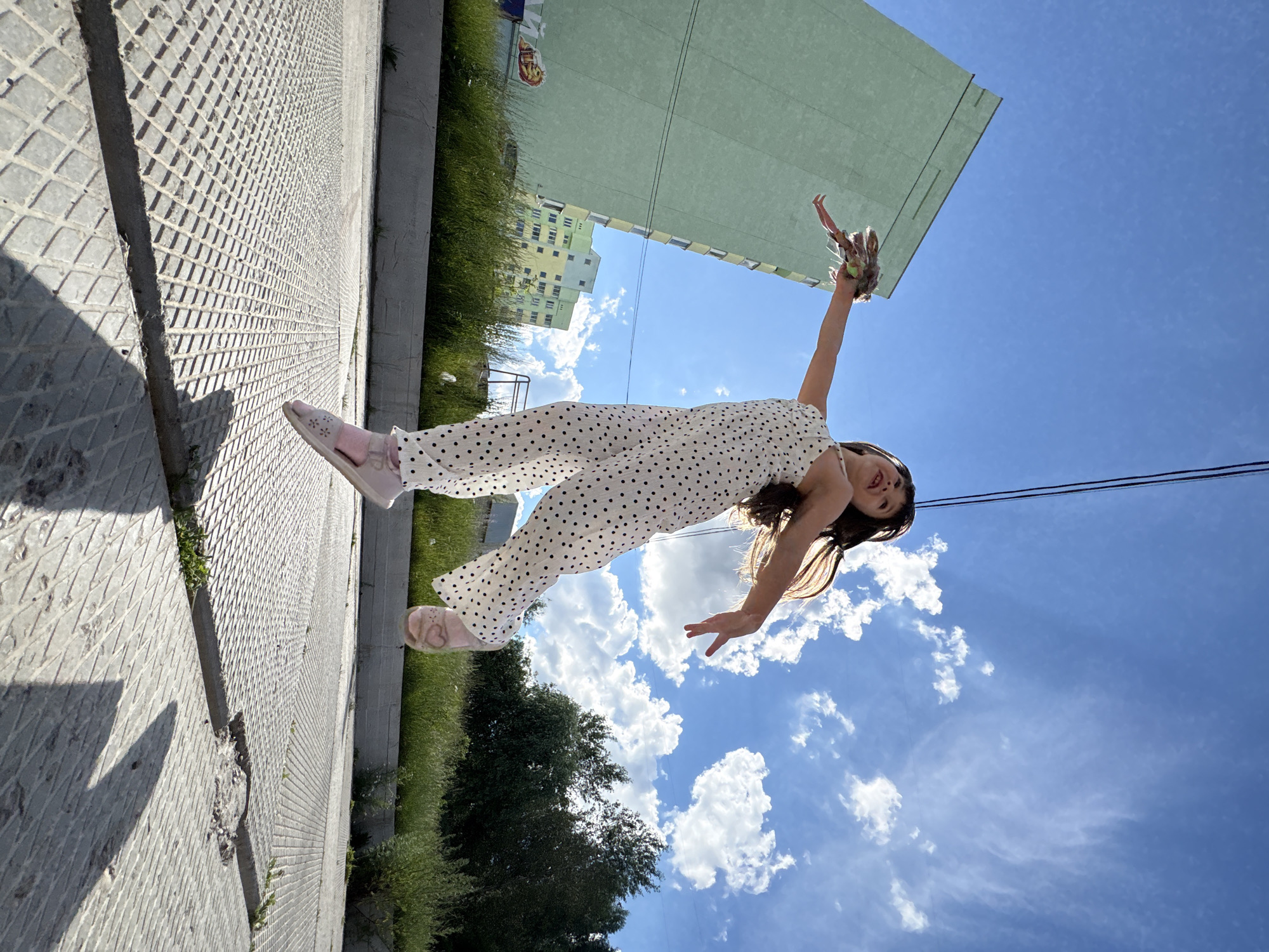 A young girl in a white polka-dot jumpsuit is captured mid-air, jumping on a concrete surface in Chaikovsky, Russia. She holds a doll in her right hand, which is extended outward. The girl's left arm is bent at the elbow, with her hand reaching forward. She is wearing pink sandals. In the background, a tall, light green building stands prominently, with a smaller, multi-colored building to its right. The sky above is a vibrant blue, dotted with fluffy white clouds. Green grass and trees form a natural boundary in the mid-ground. The concrete ground is textured with a diamond pattern. A shadow of the girl is cast on the ground. Two black wires are visible stretching across the top of the frame against the blue sky.
