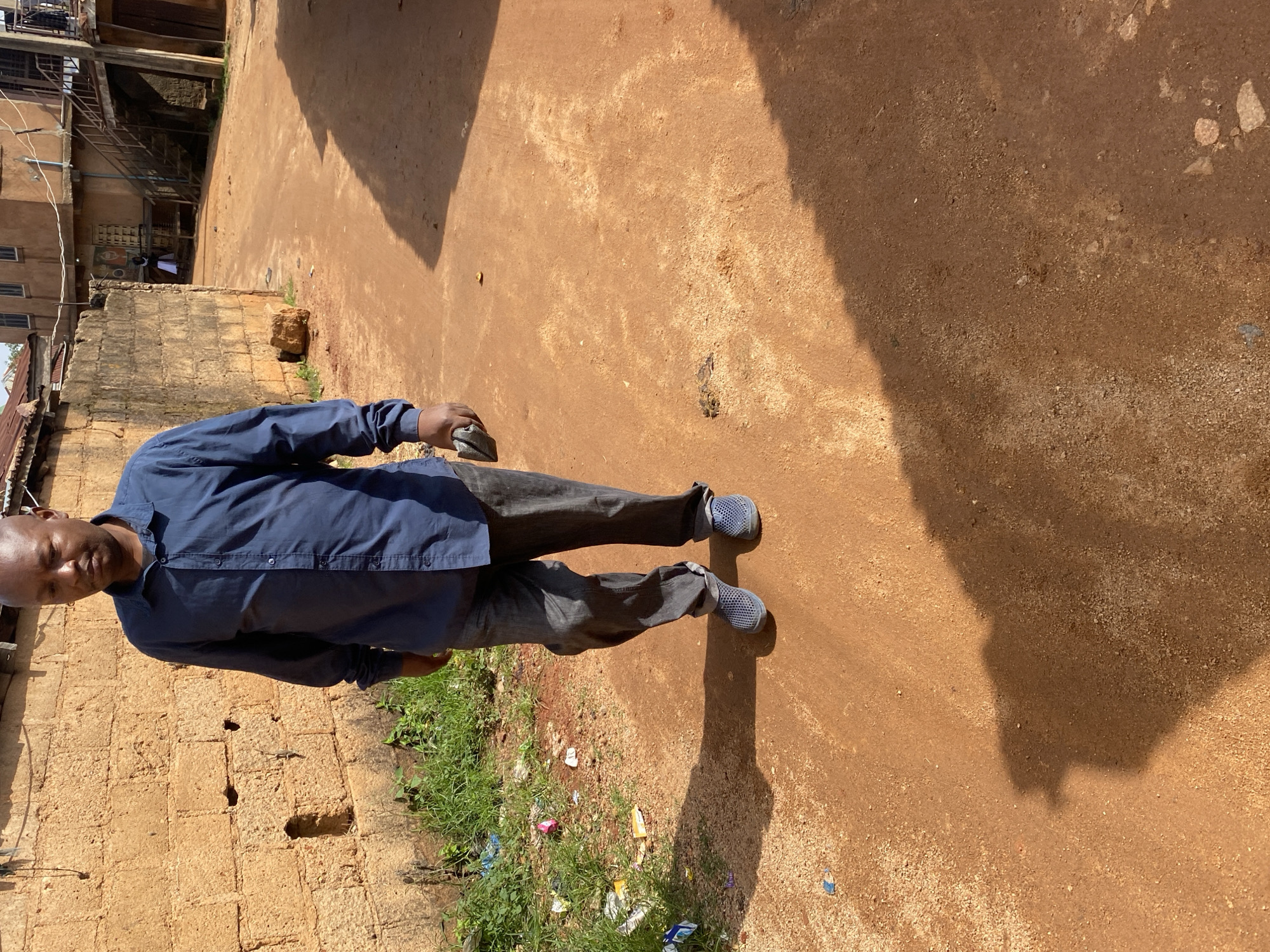 A man is walking on a dirt road in a city environment, likely in Jos, Nigeria, as indicated by the architecture and the red earth. He is wearing a blue collared shirt and dark grey jeans. His shoes are blue and appear to be made of a mesh material. He is holding a dark grey object in his right hand. The road is unpaved and dusty, with some gravel and small stones. To the left of the road, there is a low brick wall and some sparse vegetation with scattered litter. Behind the man, buildings with multiple stories are visible, some with external staircases. Sunlight is casting a long shadow of the man on the road. The overall impression is of a typical urban scene in a hot, dry climate.