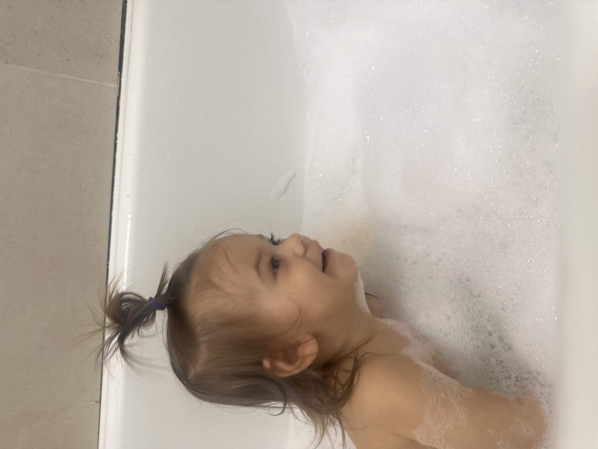 A young child is lying in a white bathtub filled with bubbles. The child has light brown hair pulled back in a small ponytail with a blue hair tie. The child is looking upwards with their mouth slightly open. The background consists of white bathtub walls and beige tiles. This image does not contain data to connect it to the city of Kharnavy, Russia.
