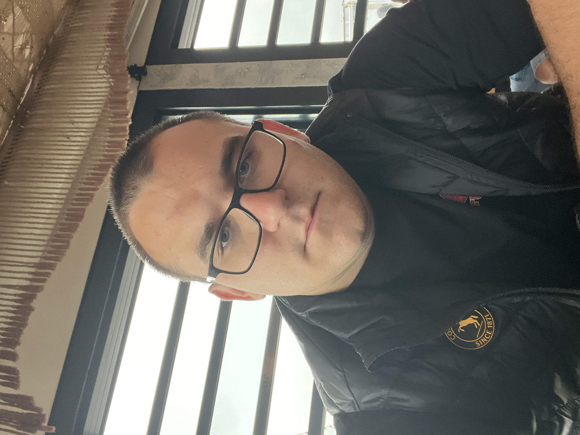 The image presents a close-up portrait of a man, taken indoors at a slight angle, likely in Varna, Bulgaria. The subject is a young to middle-aged man with short, dark hair, blue eyes, and black-rimmed glasses. His facial expression appears neutral or contemplative.
He is dressed in a black puffer vest worn over a dark t-shirt. A notable detail on his clothing is a circular gold-colored patch on the left side of his chest. This patch clearly shows the text "SINCE 1871" and features a silhouette of an animal, possibly a horse or pony, as part of a brand logo.
The background suggests an interior setting. To the left, there is a textured wall or partition with horizontal ribbed patterns, possibly made of corrugated material or woven fabric in a beige or light brown color. On the right and behind the man, large windows with dark frames are visible, allowing abundant natural light to stream in, indicating a daytime scene. The view outside is bright but indistinct, suggesting an overcast sky or bright outdoor conditions. The environment implies a public or commercial space, such as a cafe, restaurant, or a contemporary office in Varna, Bulgaria. No specific activity is taking place other than the man being photographed.