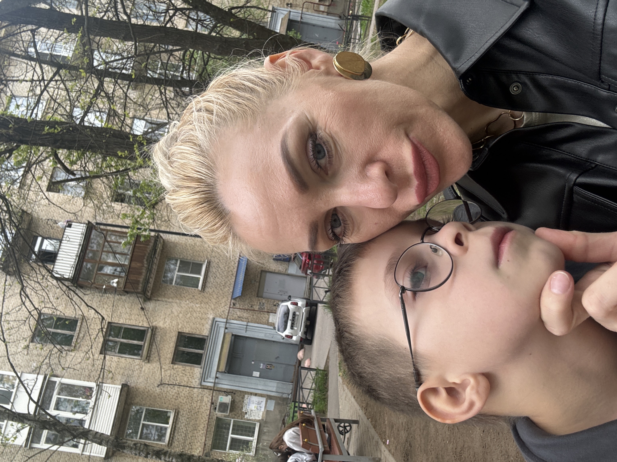 A woman with blonde hair and a black leather jacket poses with a child wearing glasses in front of an apartment building with bare trees. The location is Unknown, Unknown.