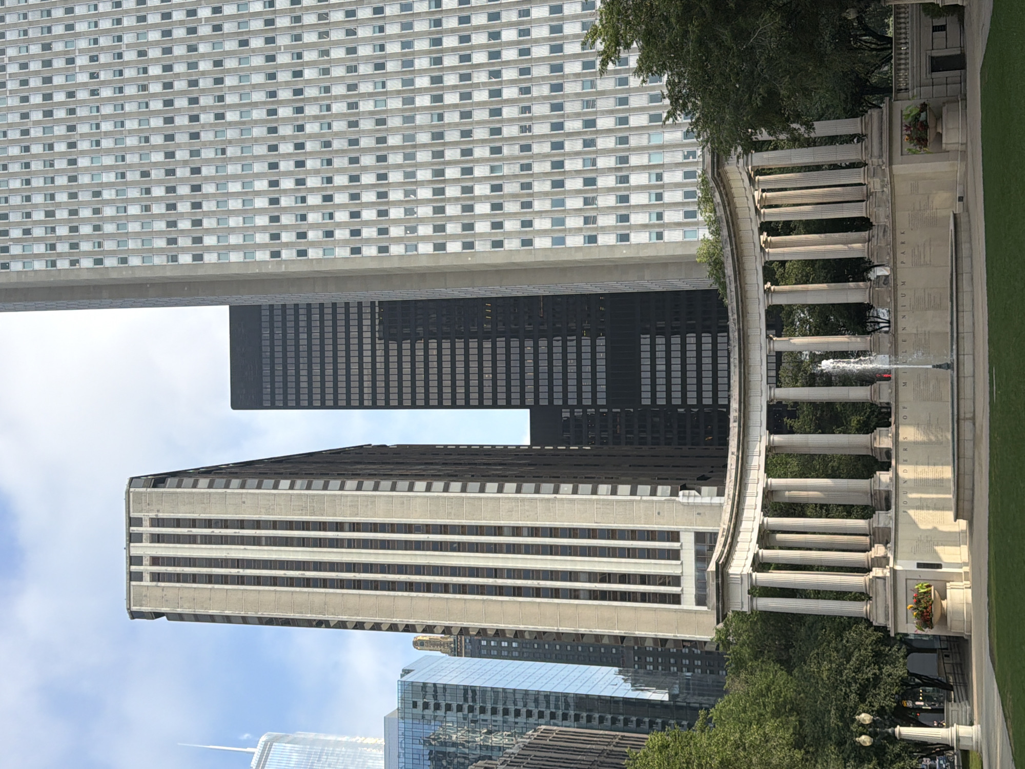 This image depicts the Jay Pritzker Pavilion and the Crown Fountain in Millennium Park, Chicago, Illinois. The scene is dominated by modern skyscrapers, including the distinctive striped facade of a tall building and a darker, grid-like skyscraper. In the foreground, a curved colonnade of white marble pillars forms a semicircular structure, likely part of the pavilion's design. A fountain sprays water in the center, adding a dynamic element to the static architecture. The surrounding area features lush green lawns and trees, suggesting a park setting on a bright, possibly overcast day, given the soft lighting. The inscription "FOUNDERS OF MILLENNIUM PARK" is visible on a plaque or wall within the colonnade. No people are visible in the frame.