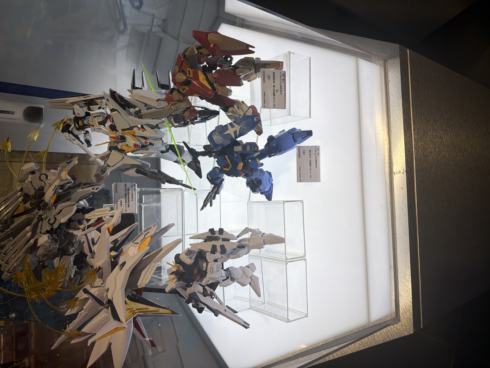 The image shows a display case with multiple Gundam model kits. In the foreground, there are two kits, one blue and one red and yellow. To the left of these, there is a white and gold Gundam model kit with sharp, angular wings. Behind this, a larger, more complex white Gundam model kit with yellow energy effects is visible. The display case is well-lit, with white light emanating from below. Information placards are visible beneath some of the models, indicating they are from Bandai Hobby. The background is dark and out of focus, suggesting an indoor environment such as a retail store. This scene is likely located in Pudong, China, a district known for its modern architecture and numerous retail establishments. There are no people visible in the image.