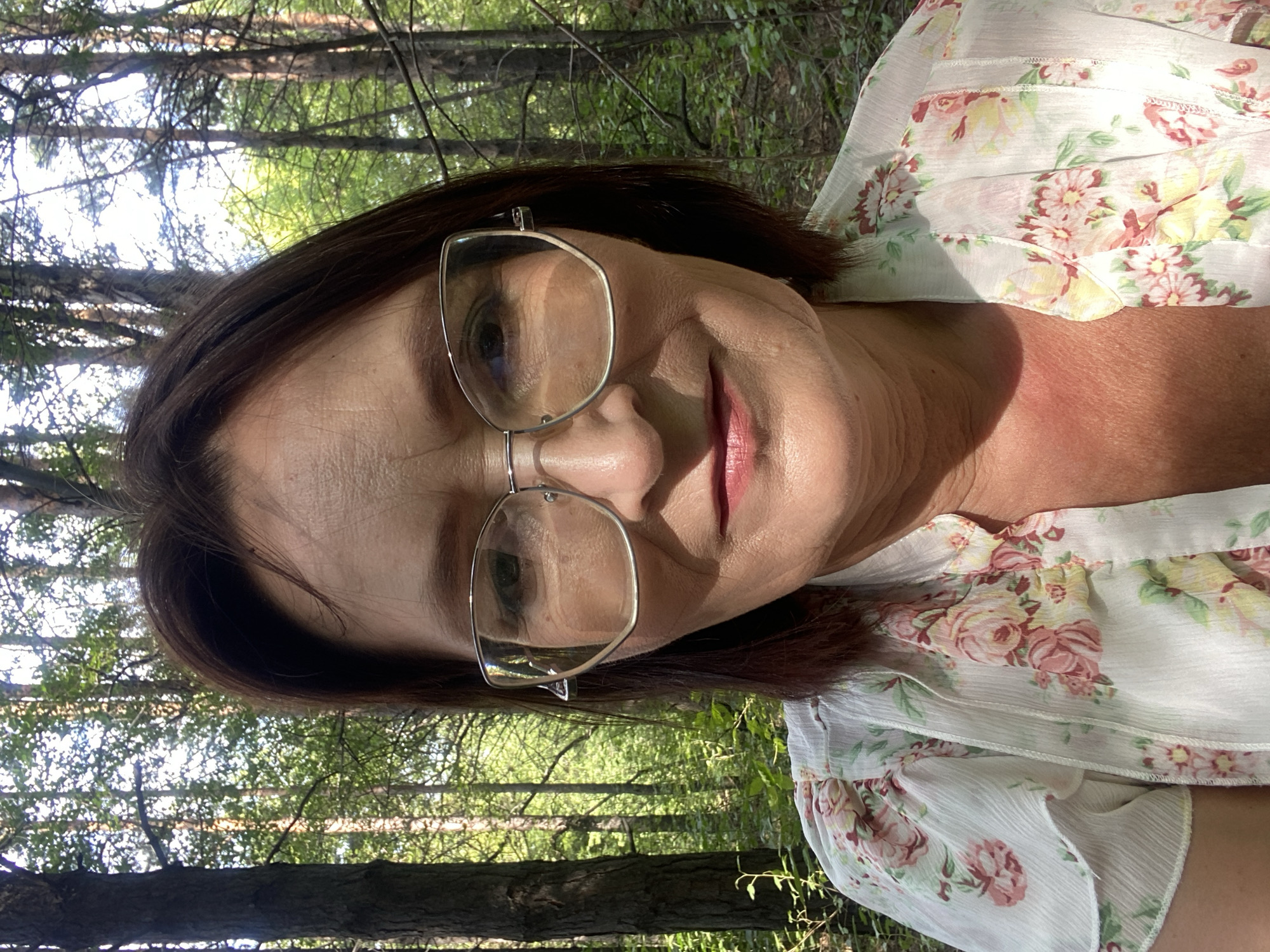 A medium shot portrait shows a woman in her late 50s or early 60s posing for a selfie outdoors. She has brown hair styled in a bob and wears large, thin-rimmed glasses. Her face is turned slightly towards the camera, and she is smiling faintly. She is wearing a light-colored blouse with a floral pattern of pink and yellow roses. The background consists of a dense forest with tall trees and dappled sunlight filtering through the leaves. The environment suggests a warm, sunny day. No specific location in Novokuznetsk, Russia is identifiable from the visual information.