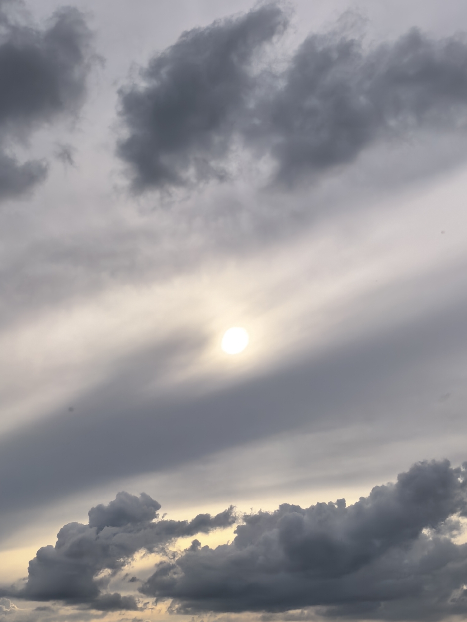 The image depicts a cloudy sky with the sun peeking through. The clouds are varied in color and texture, ranging from dark gray to lighter, wispy formations. The sun appears as a bright, luminous orb partially obscured by clouds, casting a soft glow across the sky. The overall scene suggests a dramatic or atmospheric sky, possibly before or after a storm. There are no people, structures, or identifiable landmarks present in the image, making it impossible to determine a specific location within a city or even a general environment.