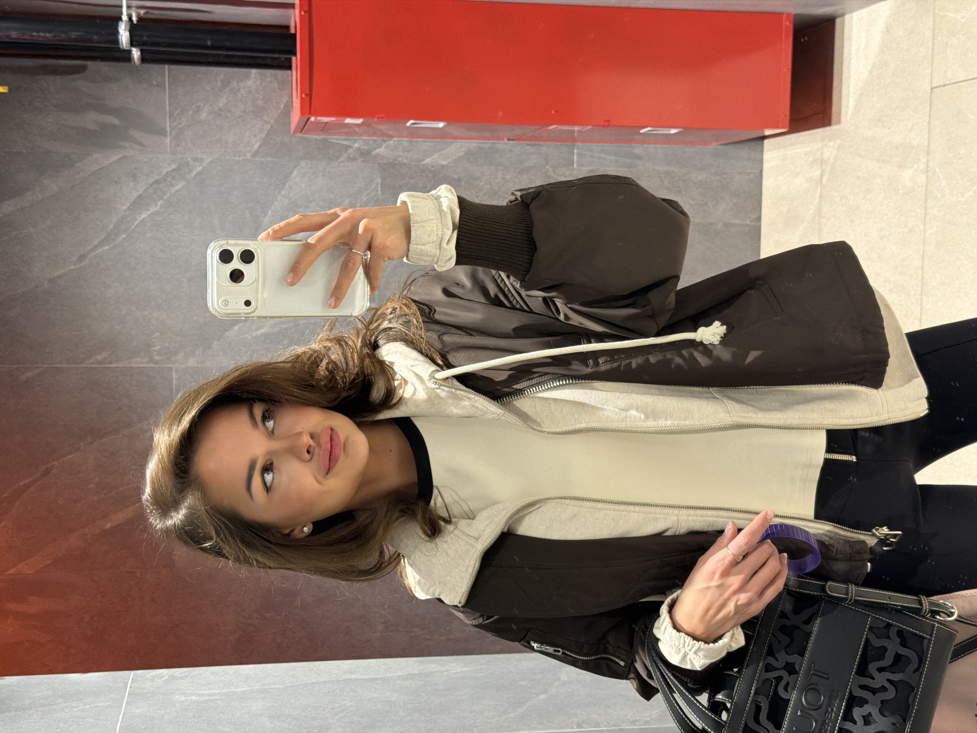 A young woman is depicted taking a selfie in an indoor setting, likely a public or commercial space in Krasnoyarsk, Russia. She holds a light-colored smartphone with a clear case in her right hand, positioned as if capturing her reflection in a mirror. Her gaze is directed towards the phone, and she exhibits a calm, pleasant expression with a subtle smile.
She has medium-length dark brown hair, styled loosely around her shoulders. Her attire consists of a layered outfit: a dark brown or black hooded jacket over a light grey hooded inner garment, and a white long-sleeve top with a dark collar. The ruffled, light-colored cuffs of the inner layer are visible. She wears a ring with a clear stone on her right ring finger and small, pearl-like stud earrings. In her left hand, which rests on a black handbag, she holds a dark purple object. The black handbag features intricate cut-out patterns, and the partial brand name "TUOT" is visible on its front.
The background shows a modern interior with large, polished grey and beige tiled flooring. A prominent red structure, possibly a cabinet or panel, stands behind her. The lighting is bright and even, suggesting artificial indoor illumination. The scene conveys a moment of self-portraiture in a contemporary, well-maintained urban environment.
