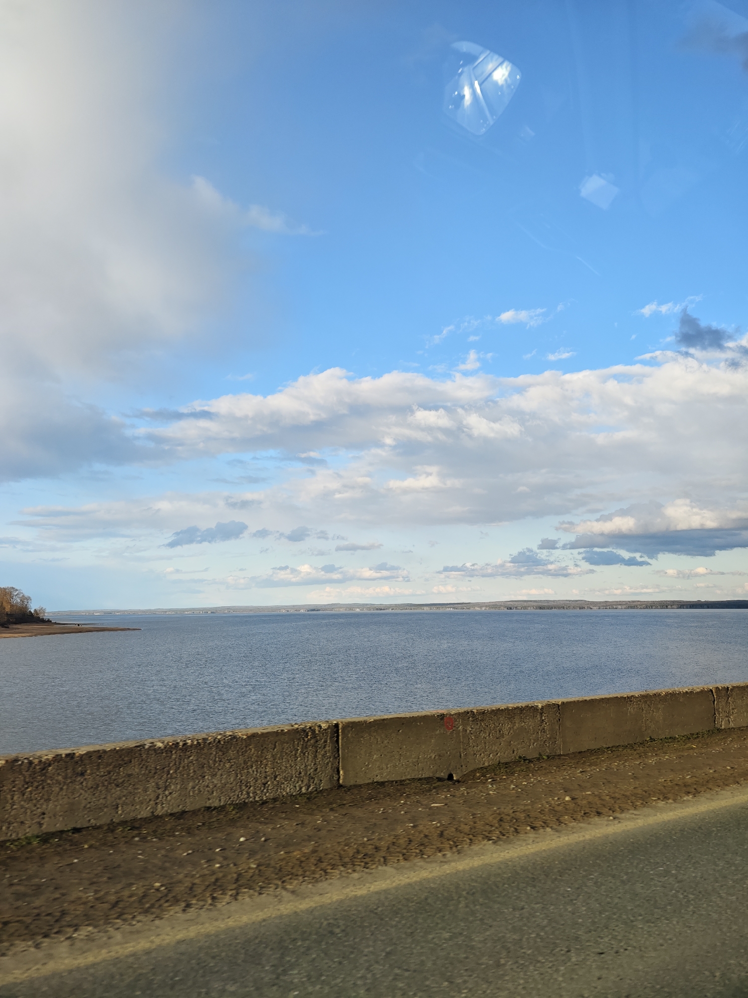 The image captures a wide, expansive view of a large body of water, likely a river or reservoir, under a dynamic sky. The scene is observed from a slightly elevated position, indicated by a concrete barrier and a section of asphalt road in the foreground, suggesting the photo was taken from a vehicle on a bridge or embankment.
The sky occupies the upper two-thirds of the frame, showcasing a mix of bright blue patches and numerous cumulus clouds ranging from fluffy white to darker grey, indicating partly cloudy weather. Reflections, possibly from a vehicle's interior like a dashboard or sunroof, are visible in the upper center of the sky, confirming the vantage point.
Below the sky, a vast expanse of calm, grey-blue water stretches towards a distant horizon. On the far left, a small, lighter-colored sandy or muddy shoreline is visible, flanked by a few bare trees, hinting at a colder season such as late autumn or early spring. Two small, indistinct figures, possibly people, can be seen on this distant shore. Across the water, a faint, low-lying landmass with a subtle line of vegetation or structures forms the horizon.
The foreground features a weathered, grey concrete barrier running horizontally across the lower part of the image, with a dirt and gravel shoulder immediately behind it. A strip of asphalt road occupies the bottom of the frame, showing signs of use.
No discernible human activity beyond the possible distant figures is taking place, and no specific event is apparent. The time of day is daylight, characterized by clear visibility and natural light. No clearly readable text is visible in the image. The overall impression is one of a vast, natural landscape experienced from a journey.
