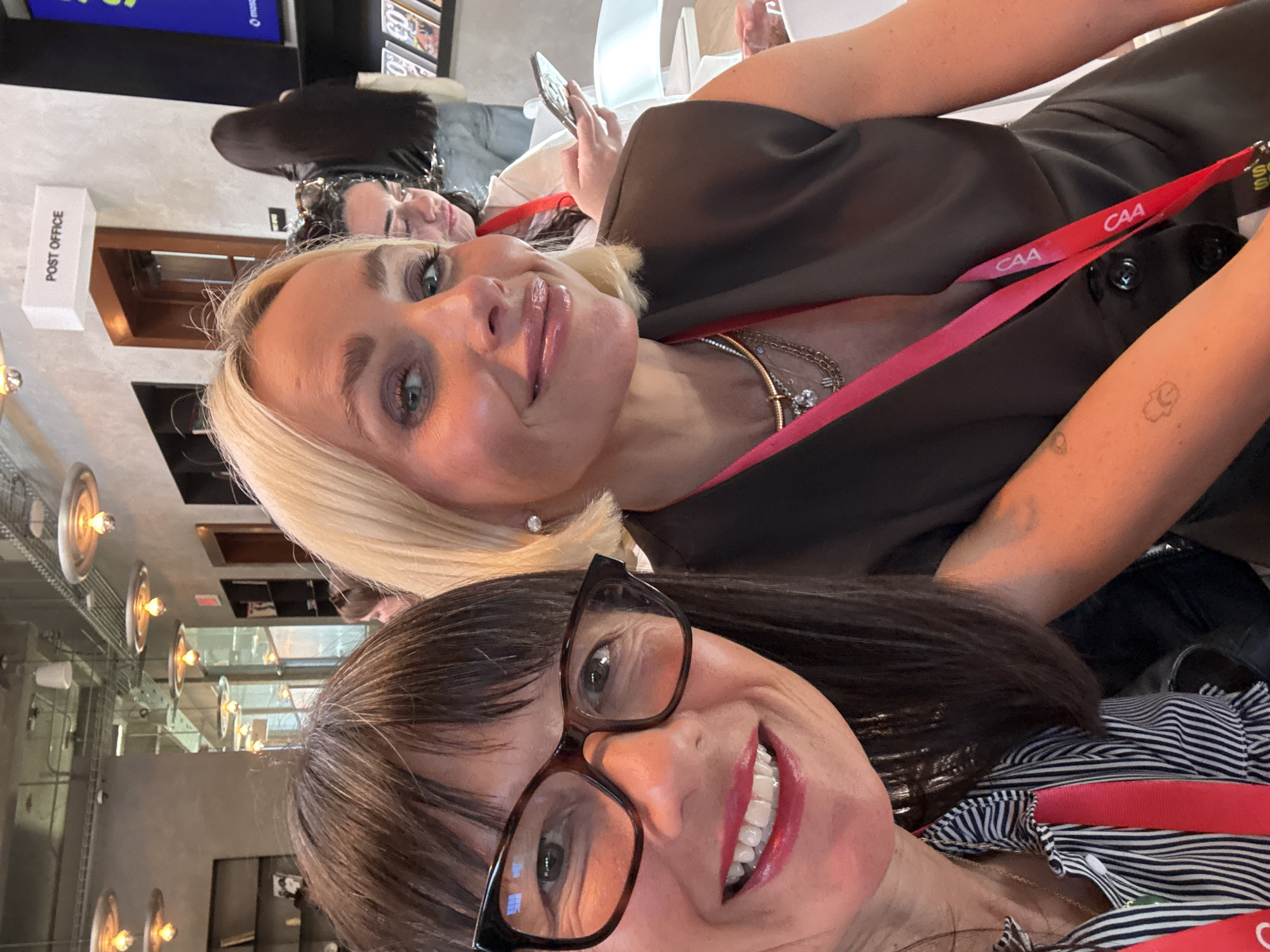 The image shows two smiling women taking a selfie indoors. The woman in the foreground has dark hair, glasses, and is wearing a black and white striped shirt with a red lanyard. The woman behind her has blonde hair and is wearing a dark-colored top with a red lanyard. Both lanyards have "CAA" printed on them. Behind the women, there is another woman with dark hair looking at a cell phone. In the background, there's a sign that reads "POST OFFICE." The location appears to be a modern office or event space with visible ceiling fixtures. The city is unknown.