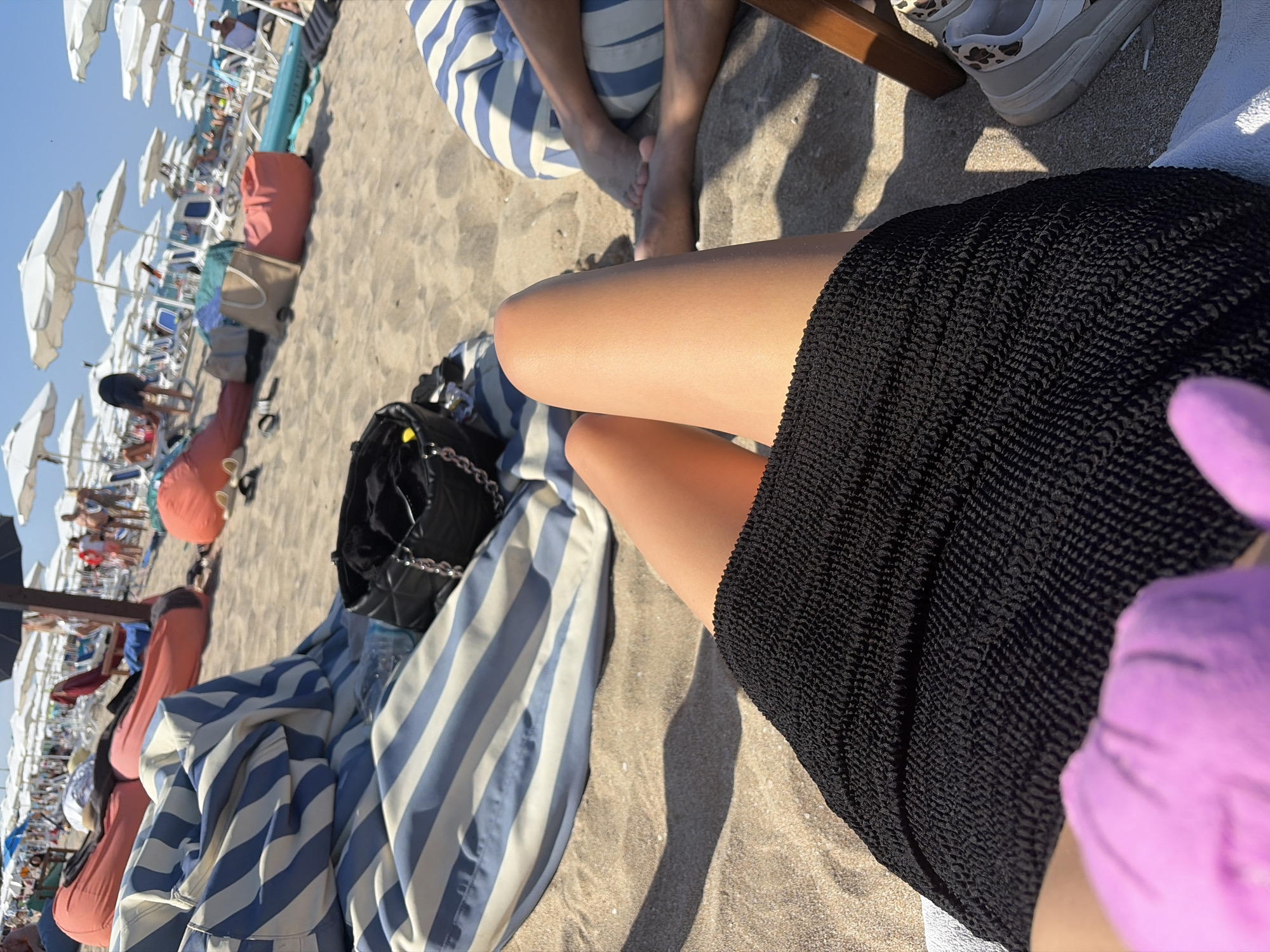 The image captures a sunny day at a beach in Lozenets, Bulgaria. In the foreground, a person's legs are visible, wearing a black textured skirt. A pink cloth, possibly a glove or fabric, is in the lower left corner. The background reveals a bustling beach scene with numerous beach chairs, umbrellas, and people enjoying the sun and sea. Several large, colorful beanbag chairs are scattered on the sand. A black quilted bag with a chain strap rests on a blue and white striped blanket. The overall atmosphere suggests a leisurely vacation setting.