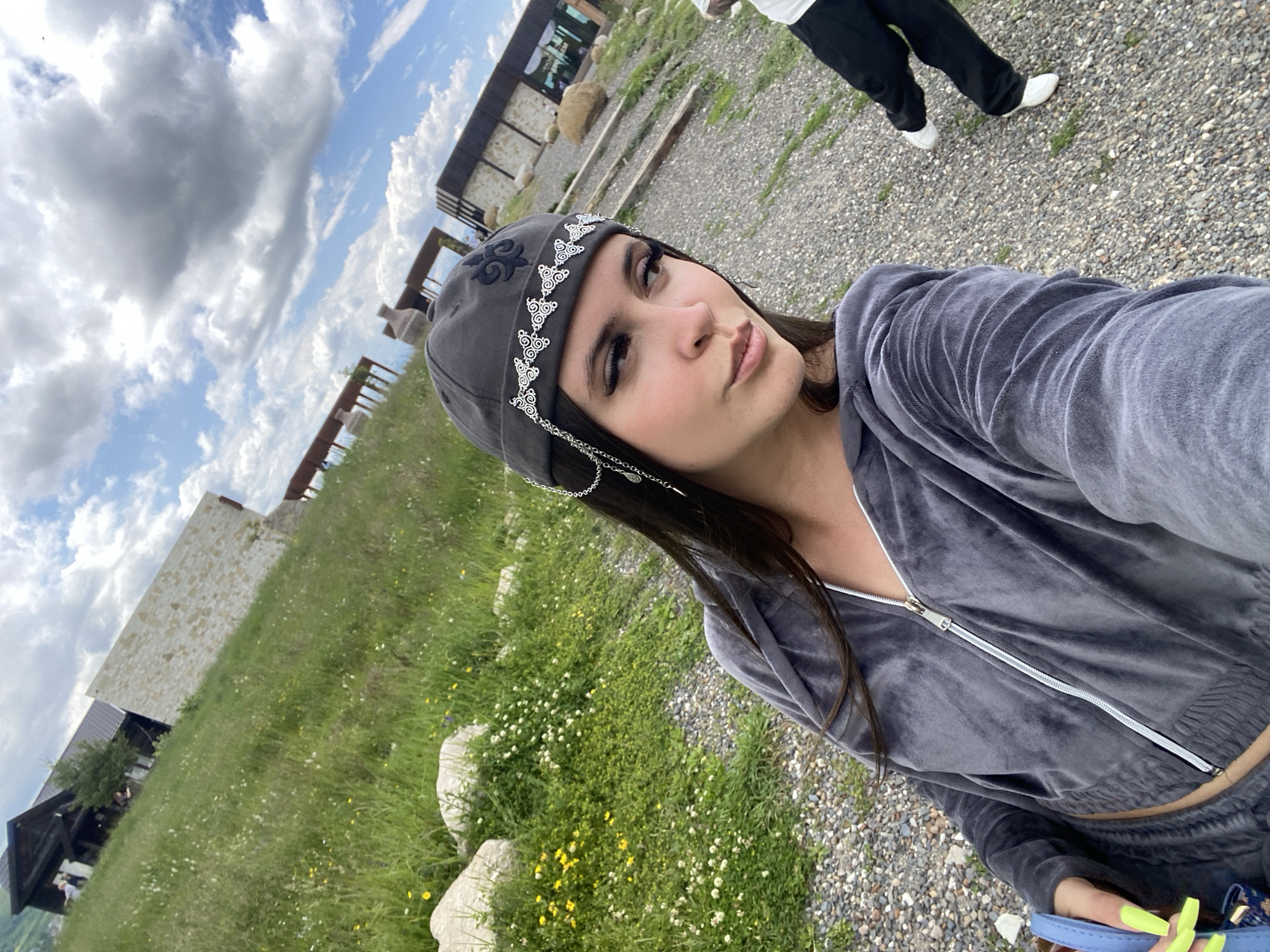 A woman is taking a selfie outdoors. She is wearing a gray velour tracksuit and a dark gray beanie with a silver chain and intricate pattern. Her left hand, with bright green nails, is visible, holding a blue bag. In the background, a gravel path leads to a modern building with a stone facade. To the left of the building, a grassy slope is dotted with wildflowers. The sky above is partly cloudy with large, dramatic cumulus clouds. There is another person walking away from the camera in the background, wearing black pants and white sneakers. The specific location is not Tsalkita, Russia; the image appears to be taken in a rural or mountainous area with contemporary architecture.
