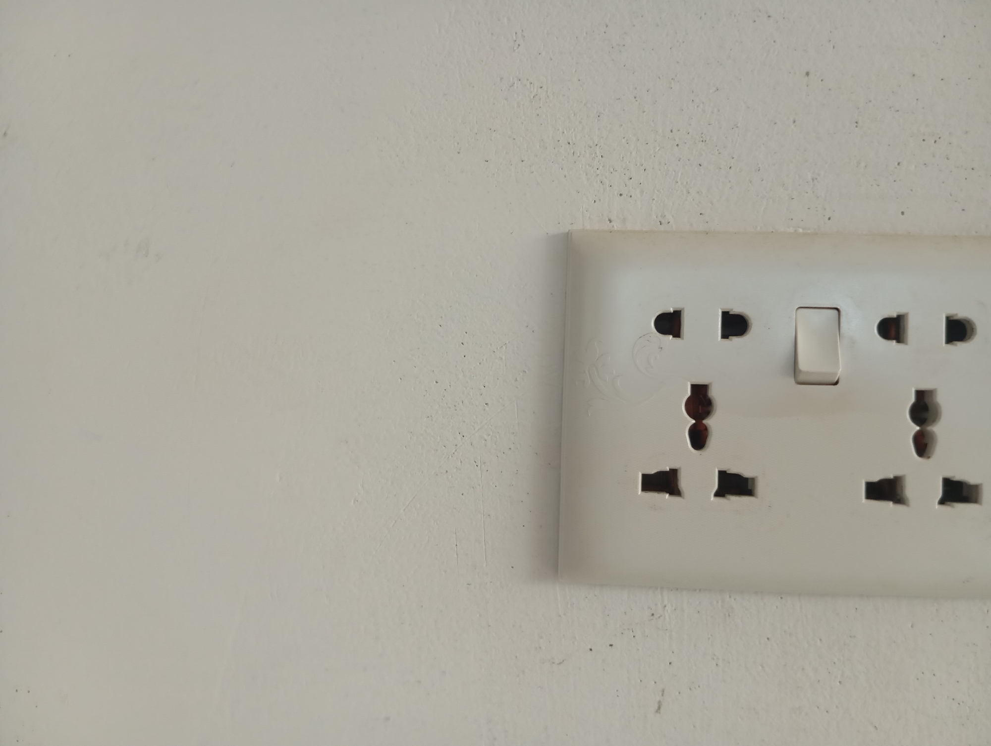 A white electrical outlet with a toggle switch is mounted on a textured white wall. The outlet has two sets of sockets, each capable of accepting multiple plug types. The switch is in the middle position. The wall is slightly uneven and shows some minor discoloration and speckling. The lighting appears to be even, casting no strong shadows. The overall scene is clean and neutral.