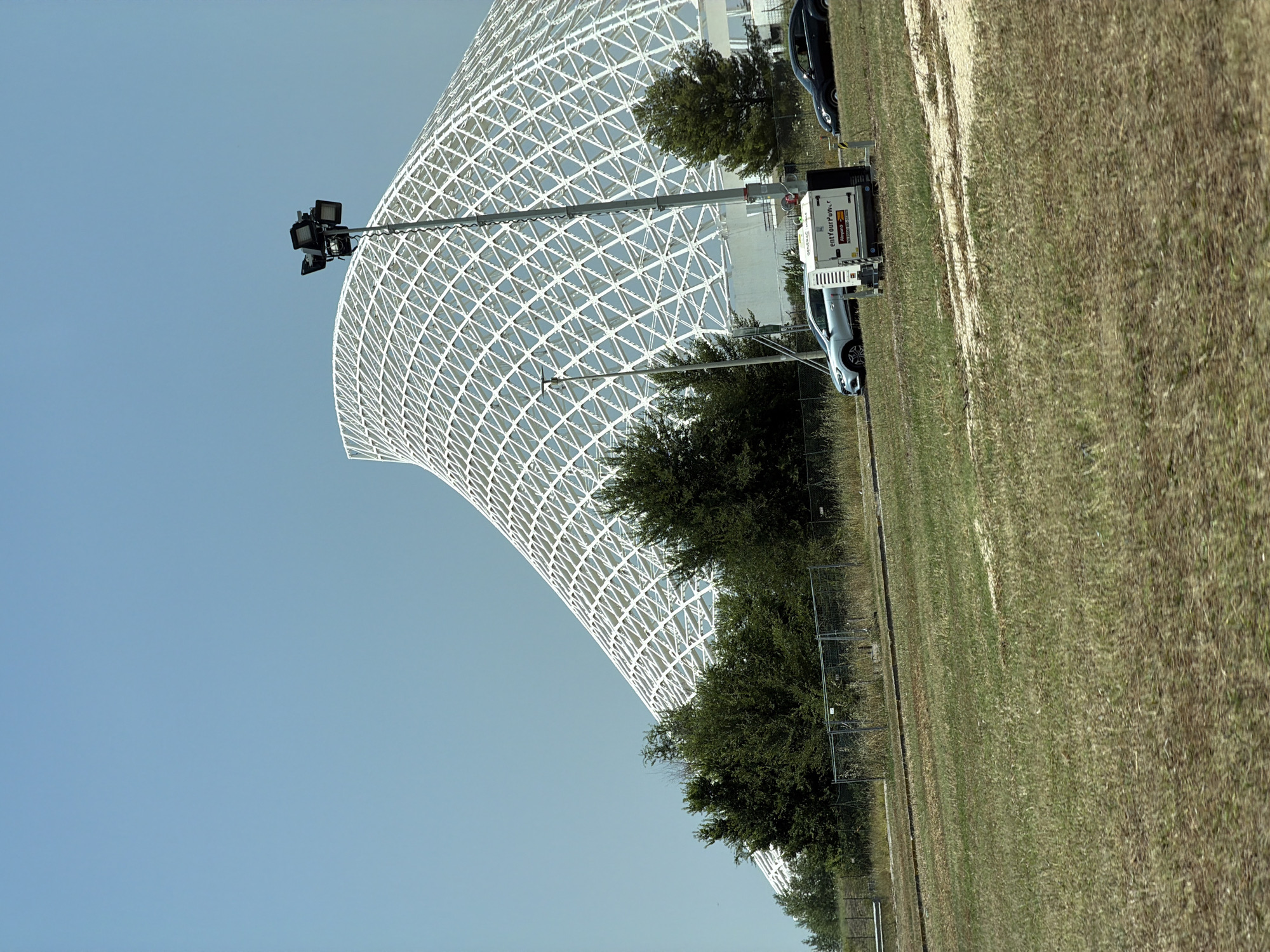 The image shows a large, white, geodesic dome structure with a lattice-like design. In the foreground, there is a grassy area and a few cars parked near a portable generator. The sky is clear and blue, suggesting it is daytime. The location is described as Unknown, Unknown.