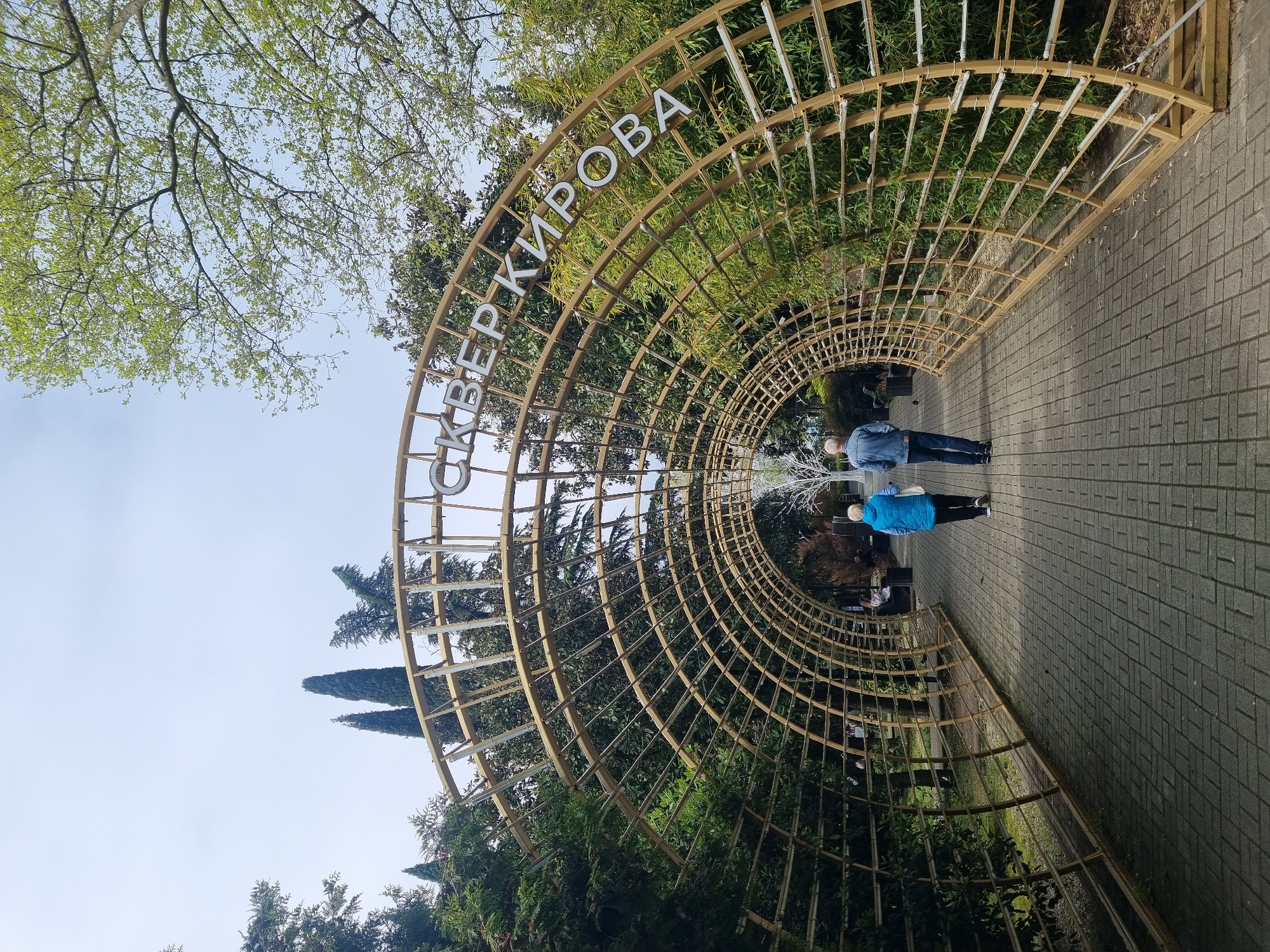 A street-level, outdoor view showcases the “Skver Kirova” in the city of Sochi, Russia. The image is rotated approximately 90 degrees clockwise.
The focal point is a semi-circular tunnel structure constructed from a grid of wooden beams. White letters spelling out "СКВЕР КИРОВА" are affixed to the top of the structure. The tunnel frames a pathway paved with gray bricks.
Two individuals are present within the tunnel's pathway. The person closest to the viewer is wearing a blue jacket and black pants. The second individual is wearing a blue shirt and darker pants. They appear to be walking away from the viewer.
Lush green foliage and trees surround the structure, with a glimpse of the sky visible above. Some of the trees, notably the cypress trees, reach tall into the sky. The light suggests it is daytime.
