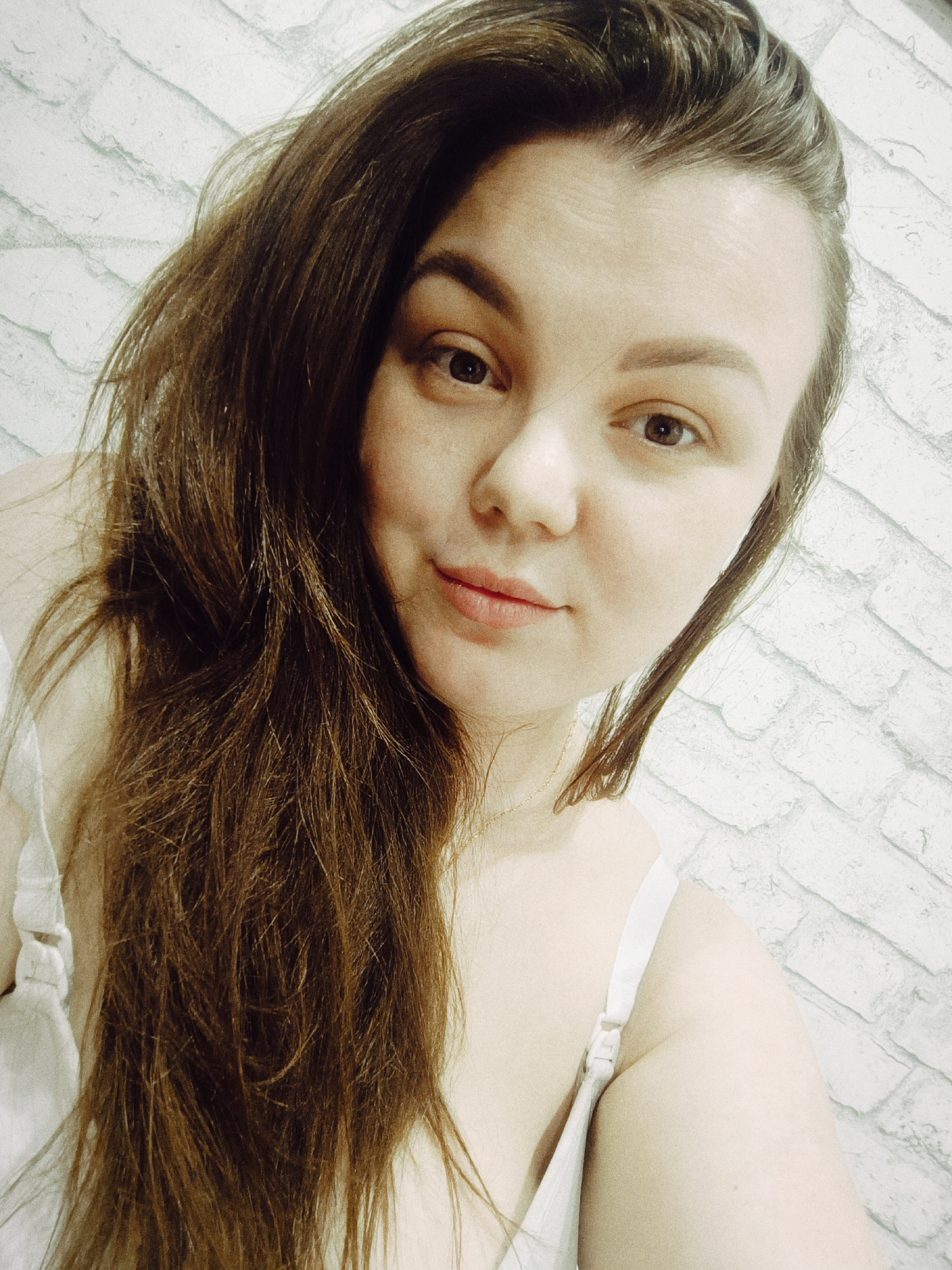 A close-up selfie depicts a woman with long, brown hair that falls across her left shoulder and down her chest. Her face is angled slightly towards the camera. The background is a textured white brick wall. The woman is wearing a white top with visible straps. A thin gold necklace is visible around her neck. The photo was taken indoors, likely in a private residence. There is no information provided about the location being in the city of Chaikovsky, Russia.