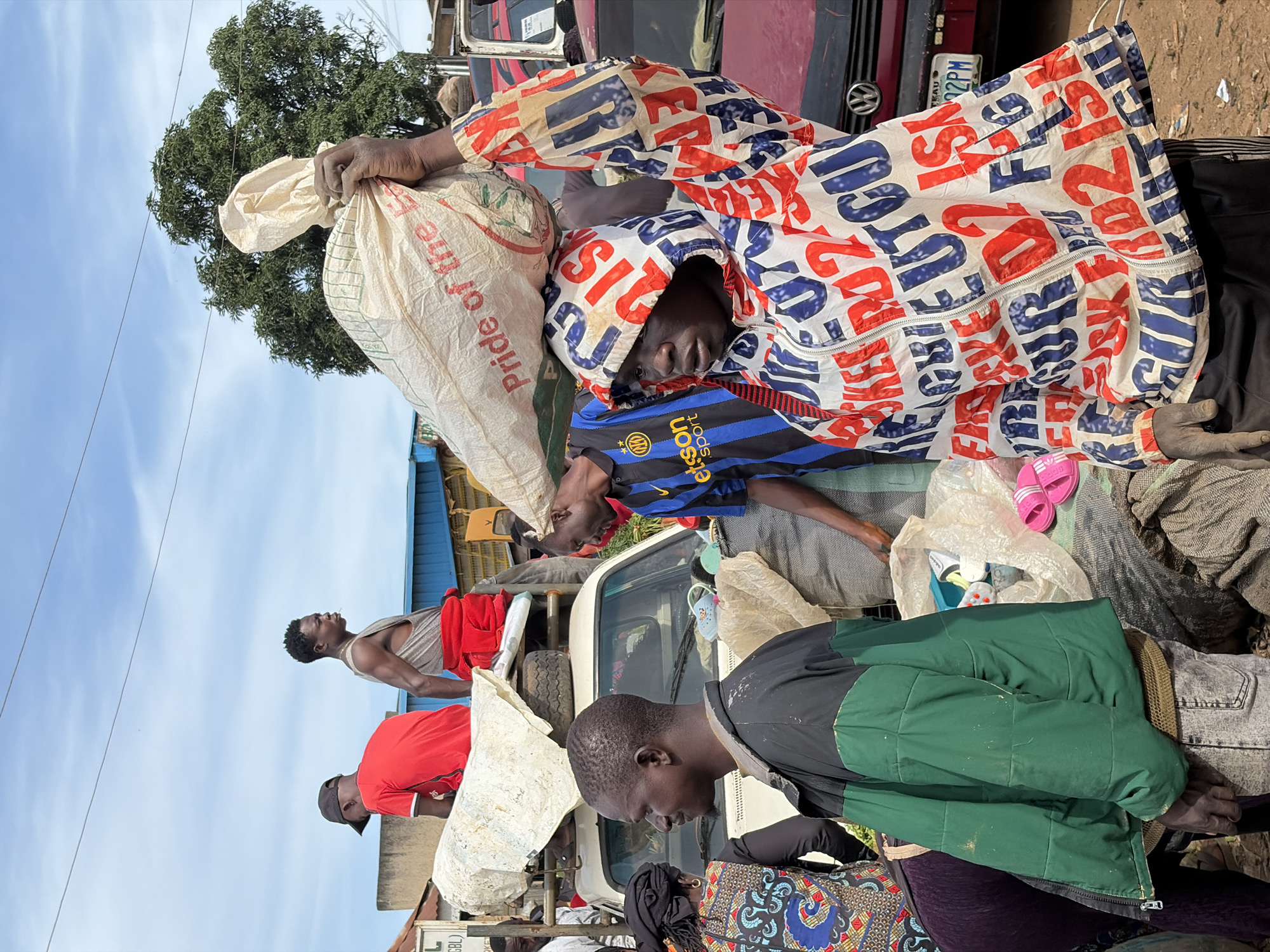 In Gura Top, Nigeria, a group of men are gathered around a truck loaded with goods. The scene appears to be a market or transportation hub. One man in the foreground is wearing a colorful jacket with bold lettering and carrying a large sack of produce on his head. Another man, in a green and black jacket, stands beside the truck. Several other individuals are visible in the background, some seated on the truck and others standing nearby. The environment suggests an outdoor setting with a clear sky and a tree in the distance. Vehicles, including a red car with a license plate, are also present. The overall atmosphere is busy and active.