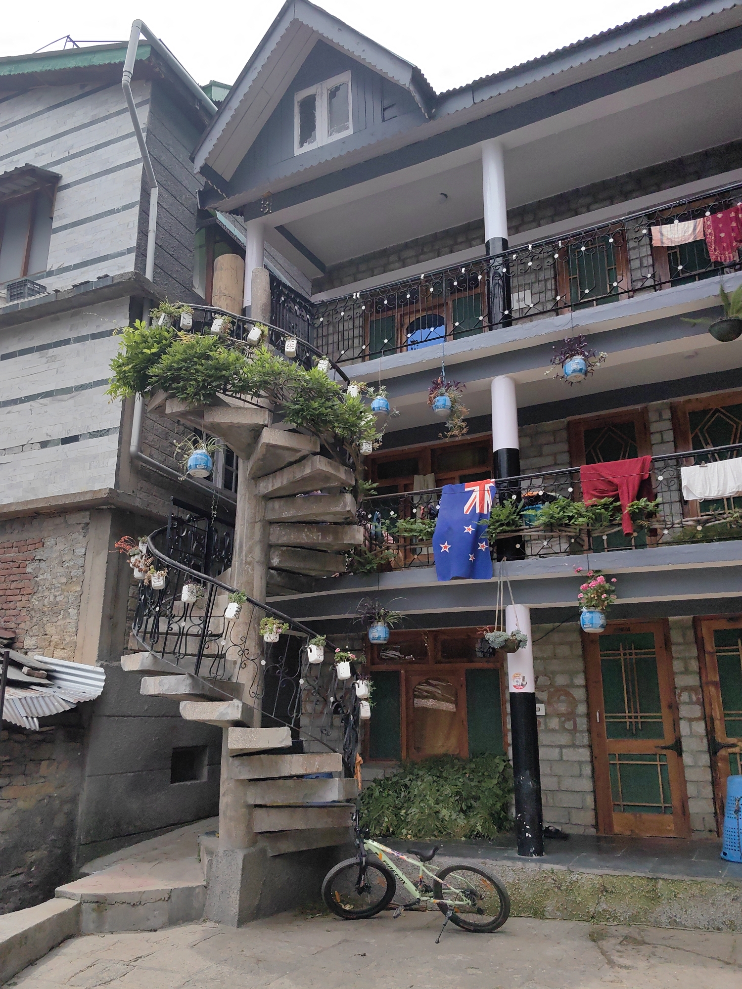 The image shows a multi-story building with a spiral staircase on the left and balconies on the upper floors. The building is made of stone and has a grey roof. There are plants on the staircase and on the balconies. A New Zealand flag is hanging on the second-floor balcony. A bicycle is parked at the bottom of the staircase. The scene appears to be in a residential area, likely in Vashisht, India, given the architectural style and the presence of greenery. The sky is overcast, suggesting a cloudy day.