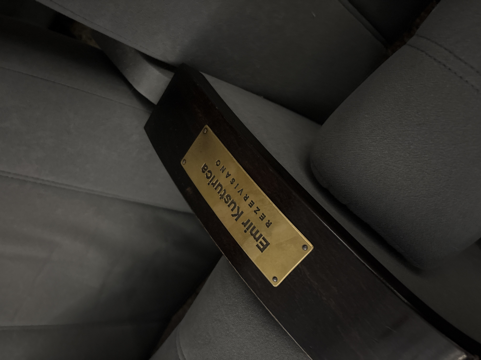 A dark-colored wooden armrest with a gold-colored plaque is attached to a gray upholstered seat. The plaque reads "Emir Kusturica Rezervisano" in black lettering. It is most likely located in Belgrade, Serbia, because Emir Kusturica is a famous Serbian film director. The word "Rezervisano" means reserved. The plaque is affixed to the wooden armrest with four silver-colored screws, one in each corner. The image is taken at a close range and the focus is on the armrest and the plaque.
