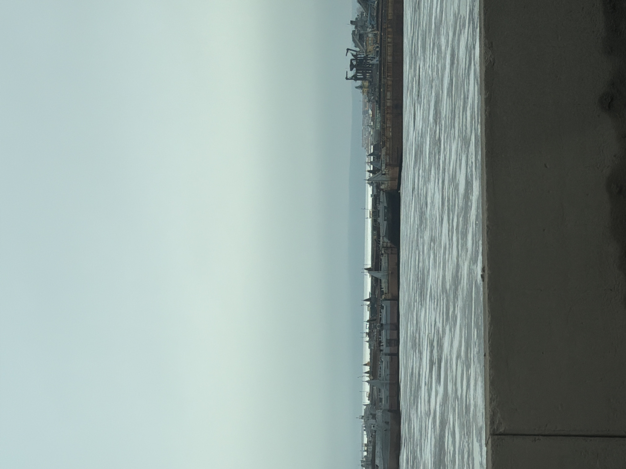 The image captures a cityscape in Chaikovsky, Russia. From a high vantage point, possibly a building, the photograph looks out over water towards a skyline marked by industrial cranes and building structures. A solid, textured structure, likely a part of the building from which the photo was taken, is visible in the foreground. The sky is overcast.