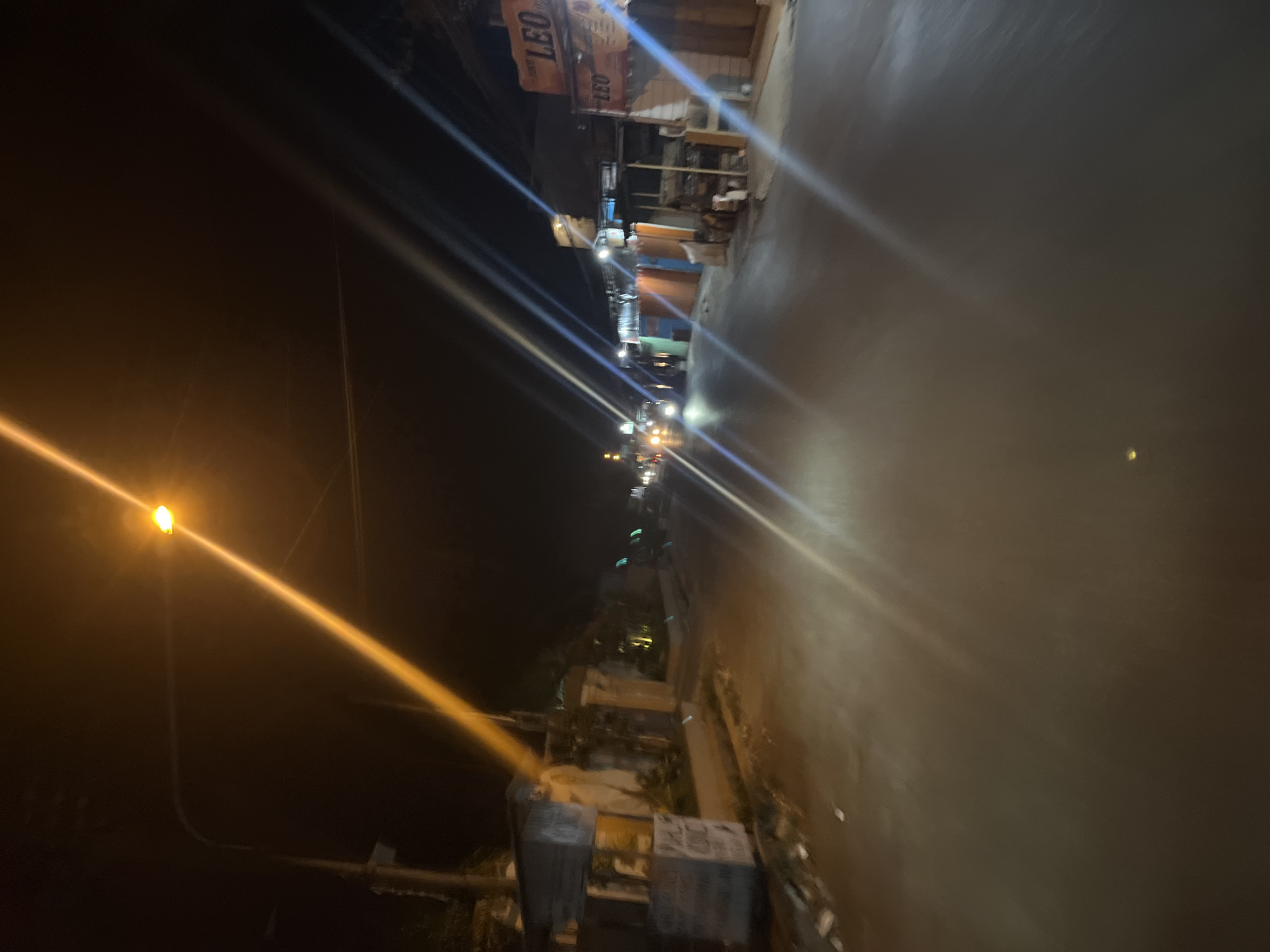 The image captures a night scene on a street in Pecangaan, Indonesia. A streetlamp illuminates the roadway, casting a bright yellow beam that diagonally traverses the frame from the upper left to the lower right. The street appears wet, reflecting the lights from streetlamps and vehicles, creating streaks of light. On the left side of the street, there are buildings and structures, some of which appear to be shops or businesses. One building has signage that reads "LEO". Several vehicles, including cars and possibly motorcycles, are visible in the distance, their headlights adding to the illuminated scene. The overall impression is of a typical urban street at night, with ongoing activity suggested by the presence of vehicles and illuminated businesses.