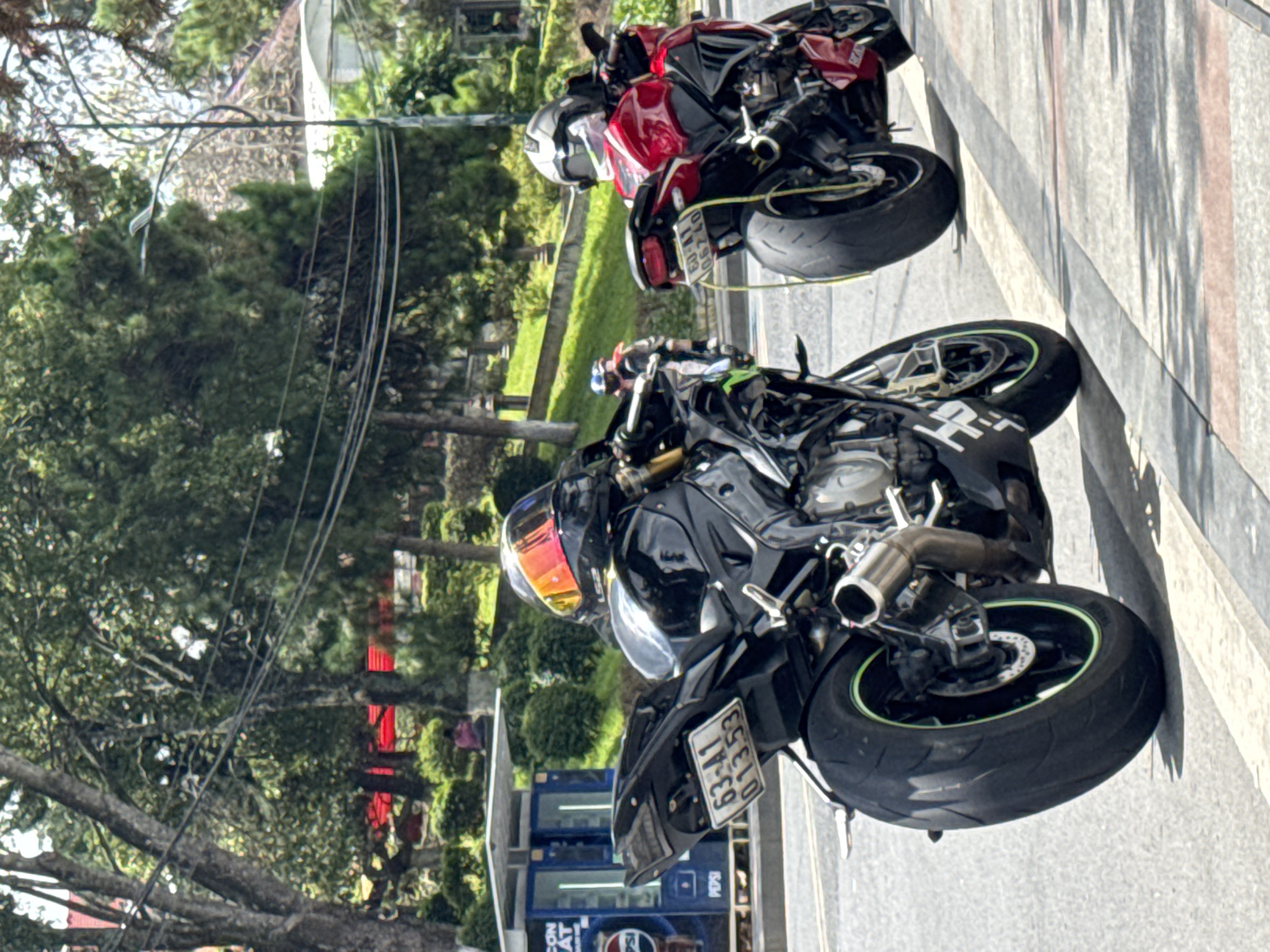 The image depicts two parked motorcycles on the side of a street in Da Lat, Vietnam. The motorcycle in the foreground is black with a reflective orange and yellow helmet on its seat. Its license plate reads "63-A1 013.53". The second motorcycle, to the right and slightly behind, is red and black with a white and grey helmet. Its license plate is visible as "60-M1 057.40". In the background, a Pepsi vending machine is partially visible, and lush green trees and foliage line the street, suggesting a park or well-maintained urban area. The lighting suggests it is daytime.