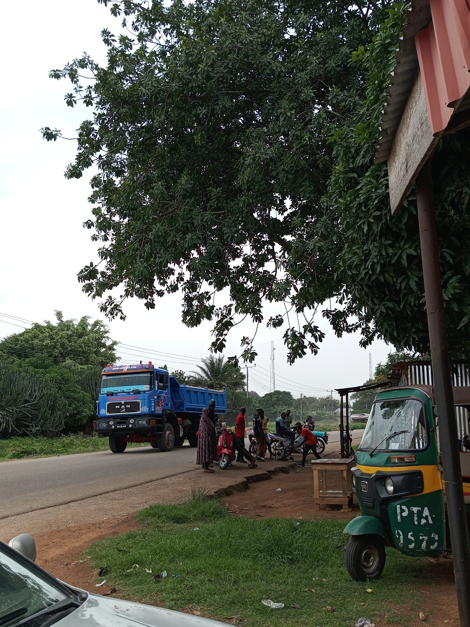 The image depicts a roadside scene in Du, Nigeria. A large blue dump truck, identified by the "MAN" logo, is parked on the left side of a paved road. Several people are gathered around the truck and on the side of the road. A woman in a patterned dress stands next to a red scooter. Further down the road, a green and yellow auto-rickshaw is parked. The background features lush green trees and vegetation, with utility poles and what appear to be communication towers in the distance. The sky is overcast. A partially visible car in the foreground suggests this is a public thoroughfare. The presence of the auto-rickshaw and the distinct style of buildings indicate an urban or semi-urban environment.